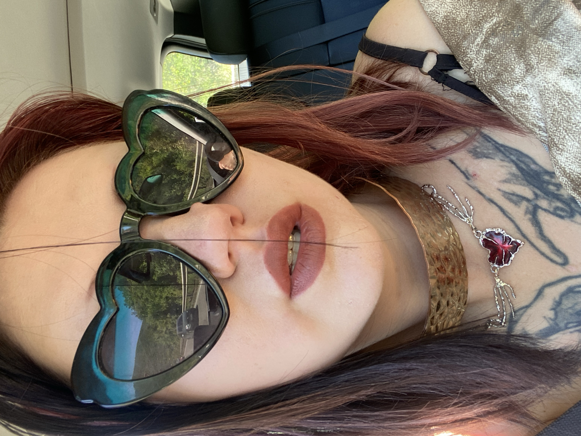 A woman with auburn hair and heart-shaped sunglasses is lying down, wearing a gold collar necklace and a silver necklace with a red pendant. She has tattoos on her neck and chest. The reflection in her sunglasses shows a street with trees and a vehicle, potentially indicating a location in Kruče - Kërruçi, Montenegro. A vehicle interior is visible in the background.