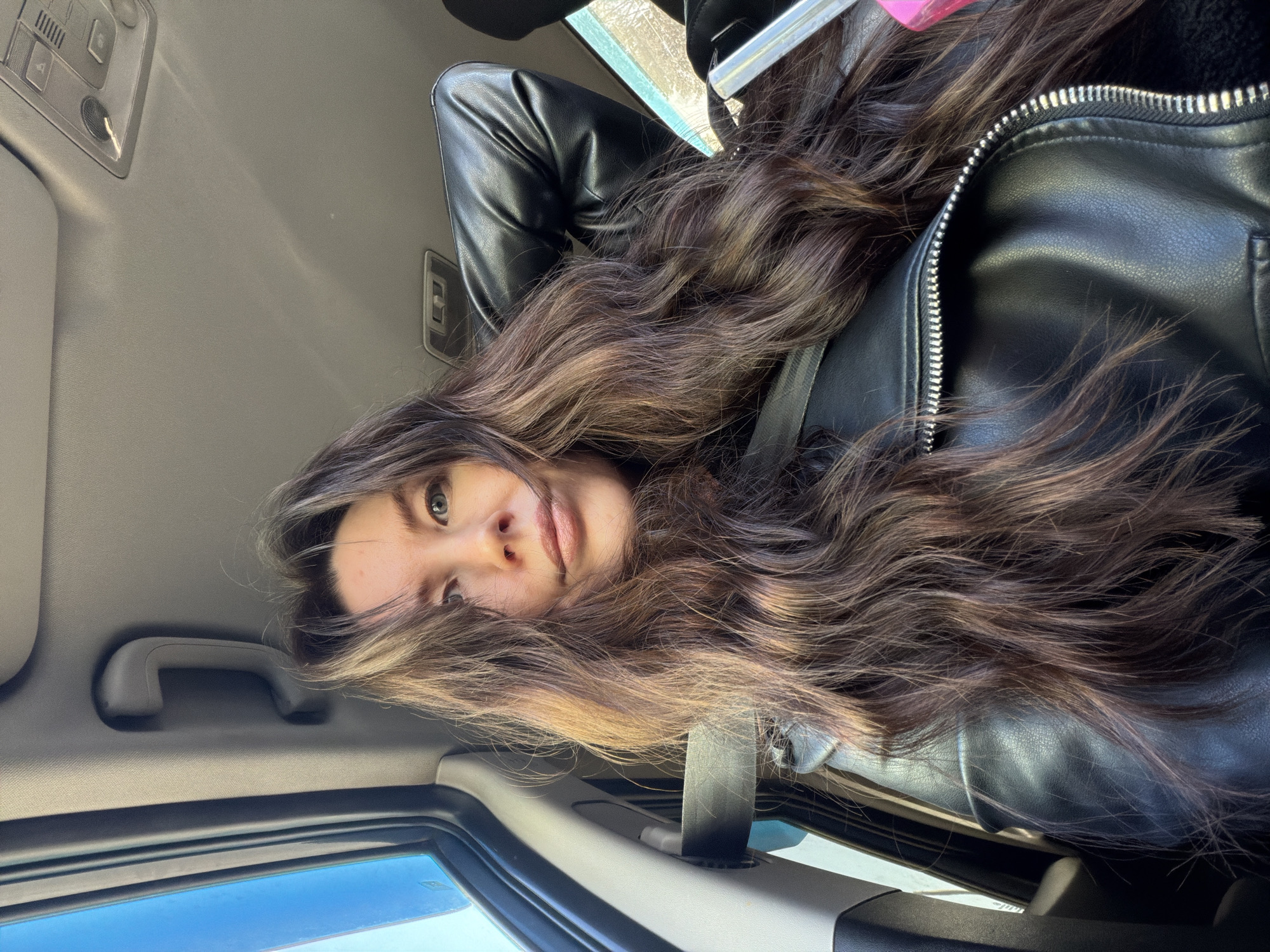 The image shows a woman inside a car, likely the passenger seat. The car's interior is visible, including the roof, grab handle, window, and sun visor. The woman has long brown hair, wearing a black leather jacket. The image is taken from an unusual angle, looking upwards. The location of the image is unknown.