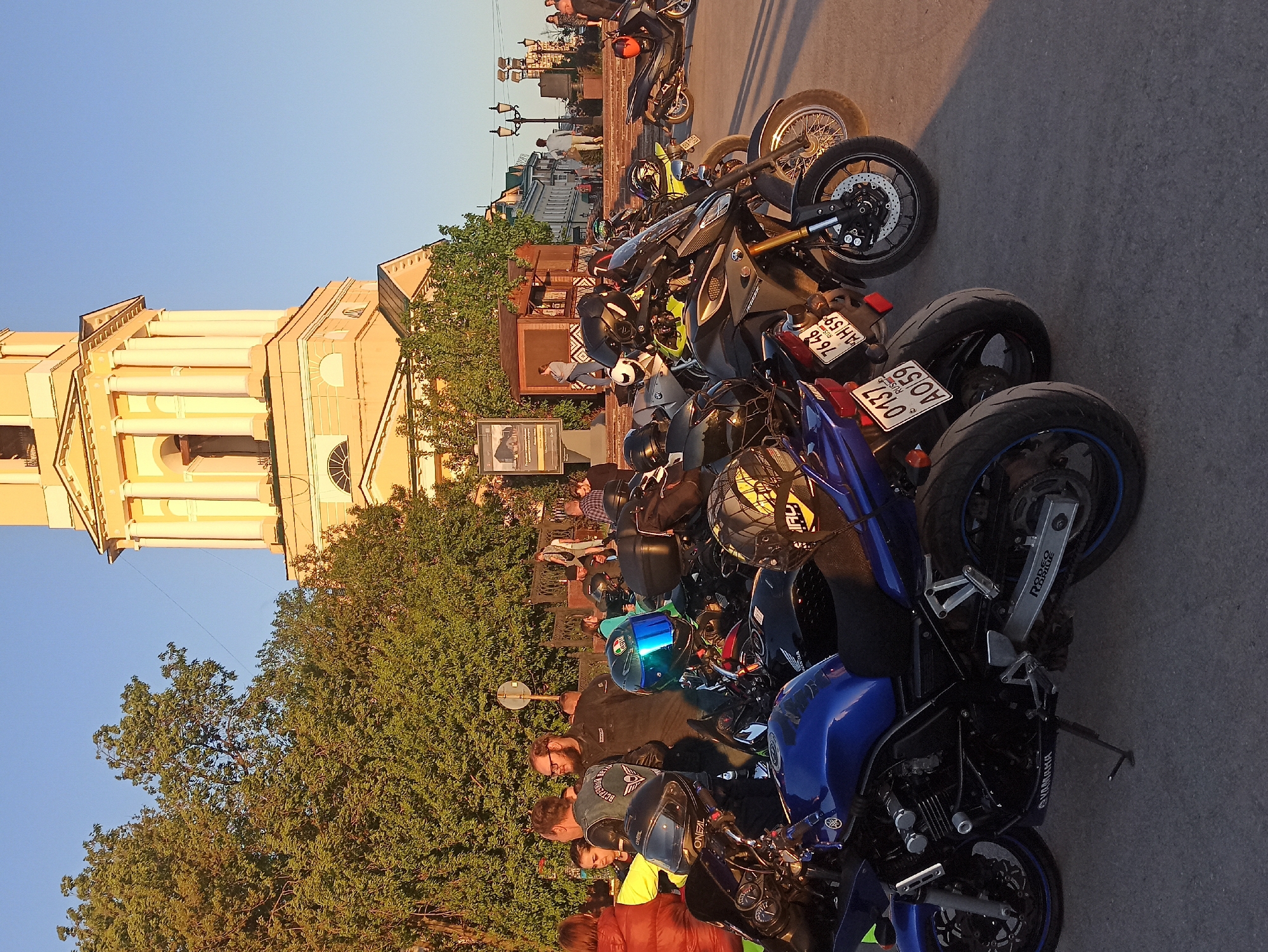 The image shows a group of motorcycles parked along a street next to a building with yellow facade. The motorcycles are various makes and models. People are gathered around the bikes. The location is Unknown, Unknown.