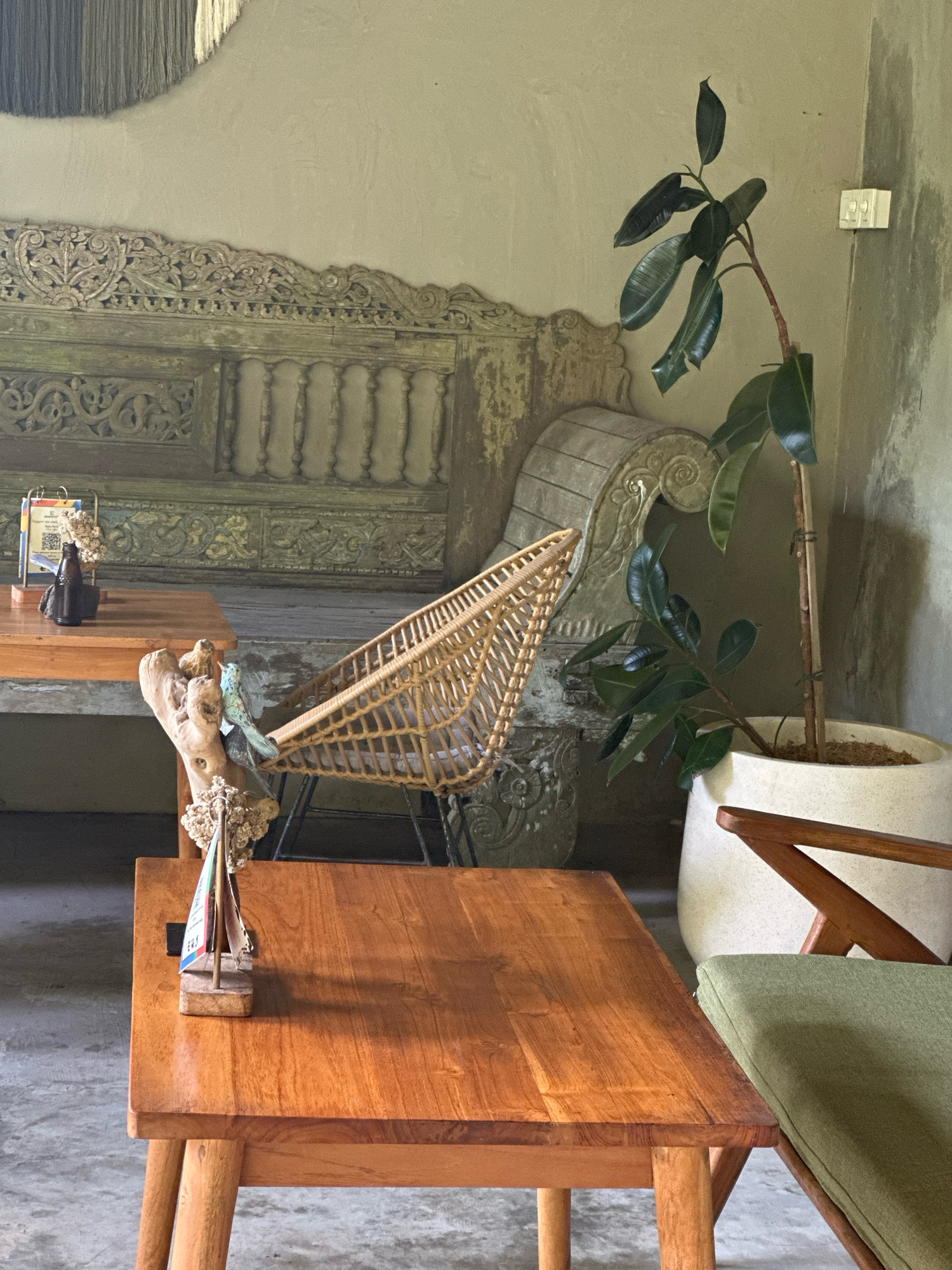 An indoor scene depicts a corner of a room, likely a cafe or lounge, with a neutral, textured wall and a concrete floor. In the foreground, a light-colored wooden coffee table with rounded edges and four legs displays a small, decorative sailboat with fabric sails on its surface. To the right of the table, a green cushioned chair with wooden armrests is visible. In the midground, a light brown woven rattan chair is positioned in front of an ornate, carved wooden bench with a distressed finish. A large potted plant with dark green, glossy leaves stands to the right of the bench. Further back, another light brown wooden table holds a menu board, a small dried flower arrangement, and a dark bottle. The overall atmosphere is rustic and serene.