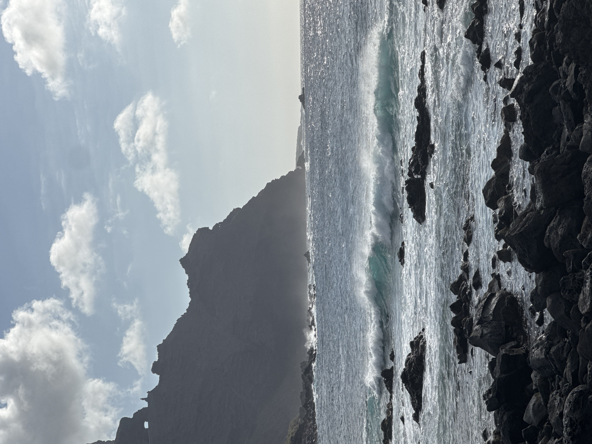 A dramatic view of a rocky coastline and the sea is presented, under a partly cloudy sky. The foreground is dominated by dark, jagged rocks and boulders, some of which are partially submerged in the water. Beyond these rocks, the sea is a deep blue, with white-capped waves crashing against the shore and breaking further out. The water has a turquoise hue in the crests of the larger waves. In the background, a massive, dark, mountainous cliff rises steeply from the sea. The cliff face is silhouetted against the bright sky, and its rugged texture is visible in places. A notable feature on the cliff is a natural archway, through which the sky is visible. Wispy white clouds are scattered across the pale blue sky, with the sun creating a bright haze in the distance. The overall scene evokes a sense of wild, untamed nature.