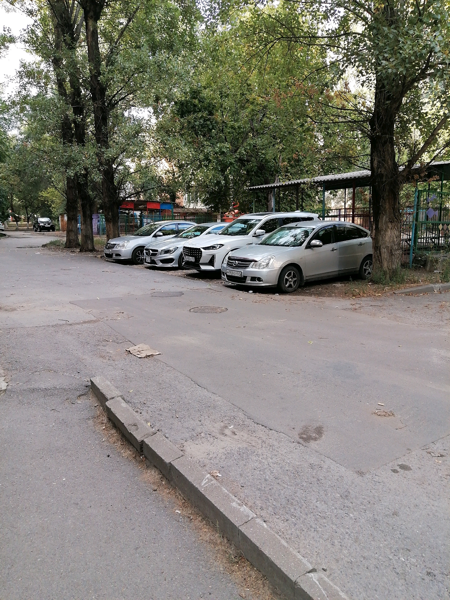 The image shows a street scene in Rostov-on-Don, Russia. Several parked cars are lined up along a paved road. The cars are of various models and colors, including silver sedans and a white SUV. Behind the cars, there are tall trees with green foliage and a low-rise building with a covered walkway. A metal fence is visible along the right side of the frame, separating the street from what appears to be a playground or recreational area with colorful structures. The road surface is worn and shows signs of repair, with some cracks and patches. The overall impression is that of a typical residential street in a moderately urban environment.