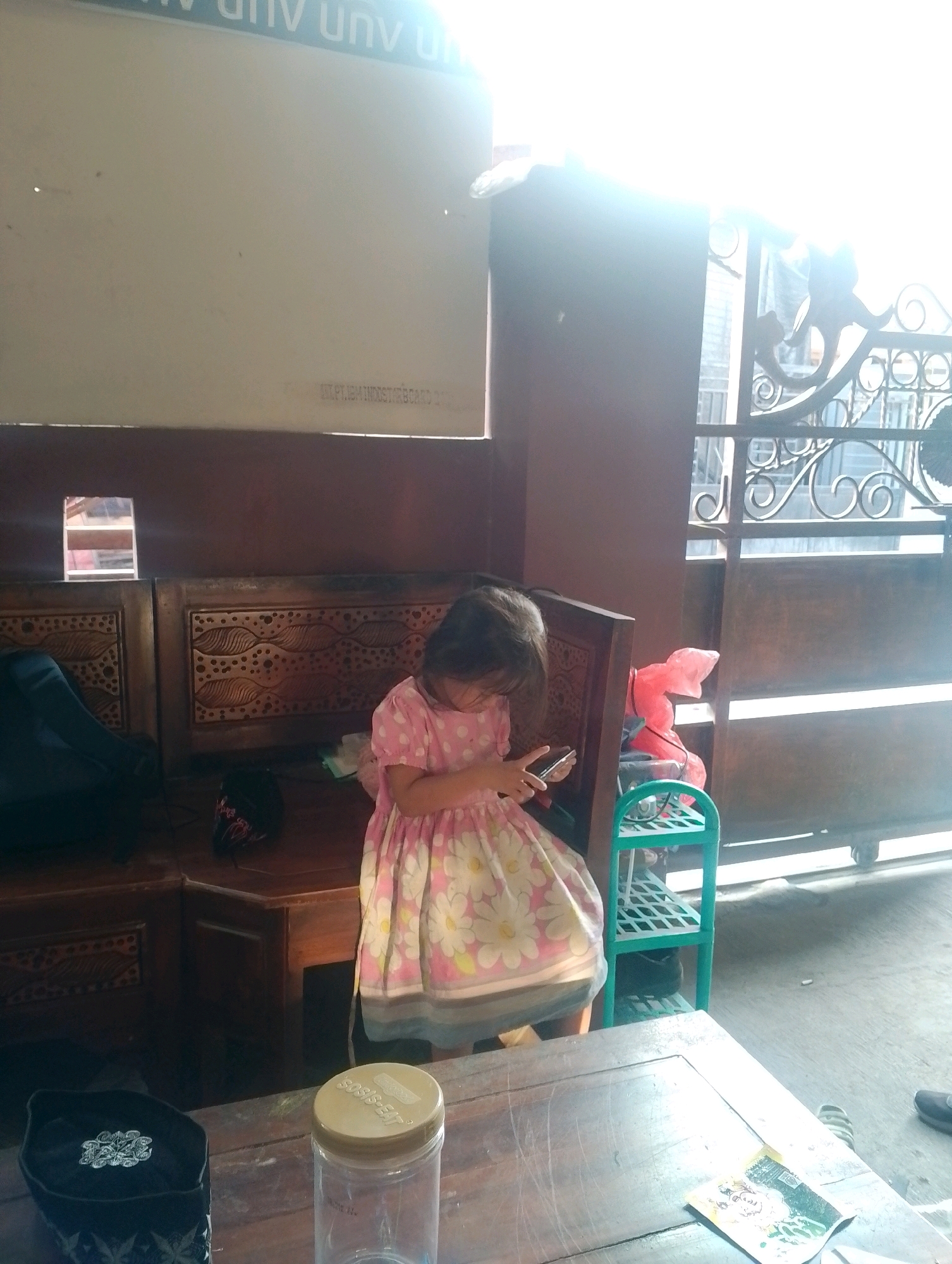 A young girl in a pink and white floral dress sits on a wooden bench, looking at her smartphone. She is in an outdoor or semi-outdoor setting, with a wooden structure behind her that appears to be part of a building or shelter. To her right, there's a green shelf unit with a pink object on it. In the background, a decorative metal gate is visible. In the foreground, a wooden table holds a black cap with intricate embroidery and a clear plastic jar with a beige lid. The scene is brightly lit, possibly by sunlight. The location is identified as Kendalrejo, Indonesia.