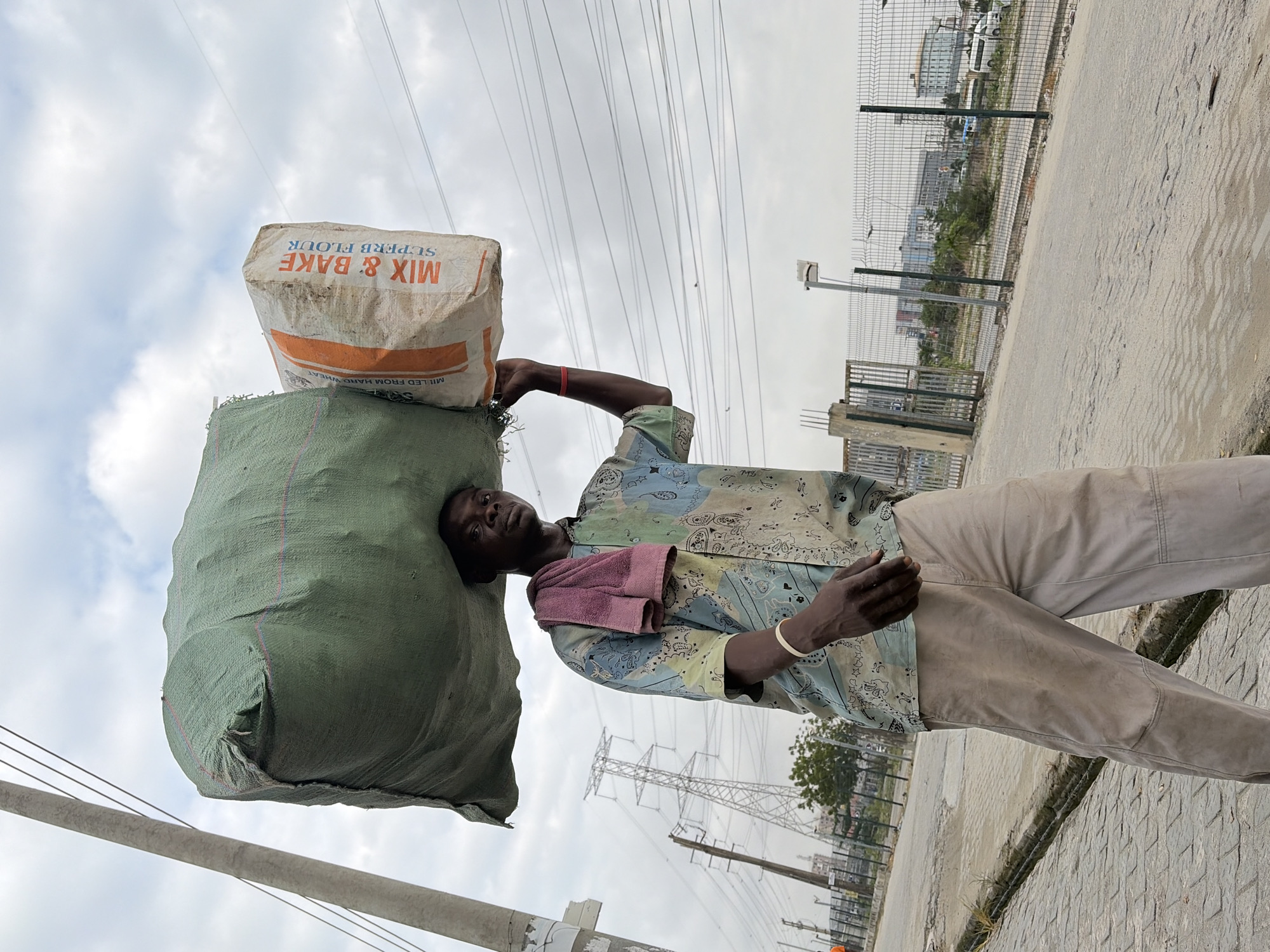 A man walks on a paved road in Igboefon, Nigeria, carrying a large green bag and a smaller white bag of flour on his head. He wears a short-sleeved, patterned shirt and light-colored pants. A pink towel is draped over his left shoulder. In the background, there are power lines, utility poles, a fence, and buildings. The sky is cloudy.