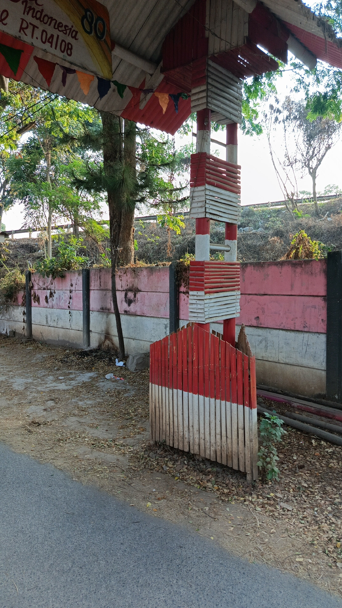 In the city of Margajaya, Indonesia, a weathered concrete wall, painted with horizontal bands of pink and white, stretches across the scene. The wall is partially obscured by a tall, slender tree with sparse foliage. In the foreground, a red and white striped pole with horizontal wooden slats rises from a small, similarly striped fence. This structure appears to be part of a larger roofed area, indicated by a corrugated metal roof overhead, adorned with a string of colorful triangular flags and a banner that reads "Indonesia" and "RT.04108". The ground is covered in dry leaves and dirt, with a section of asphalt road visible in the lower left corner. The overall impression is one of a modest, outdoor structure in a somewhat neglected, tropical environment.