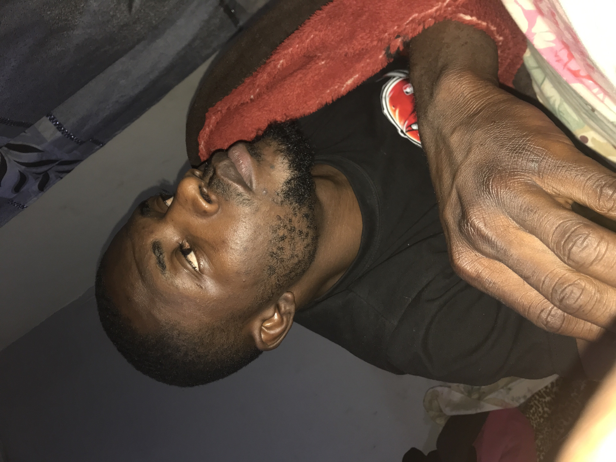 This image depicts an adult male in an indoor setting in Rafin Par, Nigeria. The individual, with dark skin, short dark hair, and a short beard, is wearing a black t-shirt with a partially visible red and white logo. He is positioned in a reclining or resting posture, looking upwards and to the viewer's left. A reddish-orange textured blanket is draped over his right side, and his right hand, showing prominent skin texture, rests in the foreground. Beneath his hand and arm, a patterned sheet or pillowcase with floral or abstract designs in pink, white, and green is visible. The background features a plain dark wall on the left and a darker, patterned curtain or fabric with leaf-like motifs and reflective embellishments on the upper right. The scene is dimly lit, suggesting an indoor domestic environment.