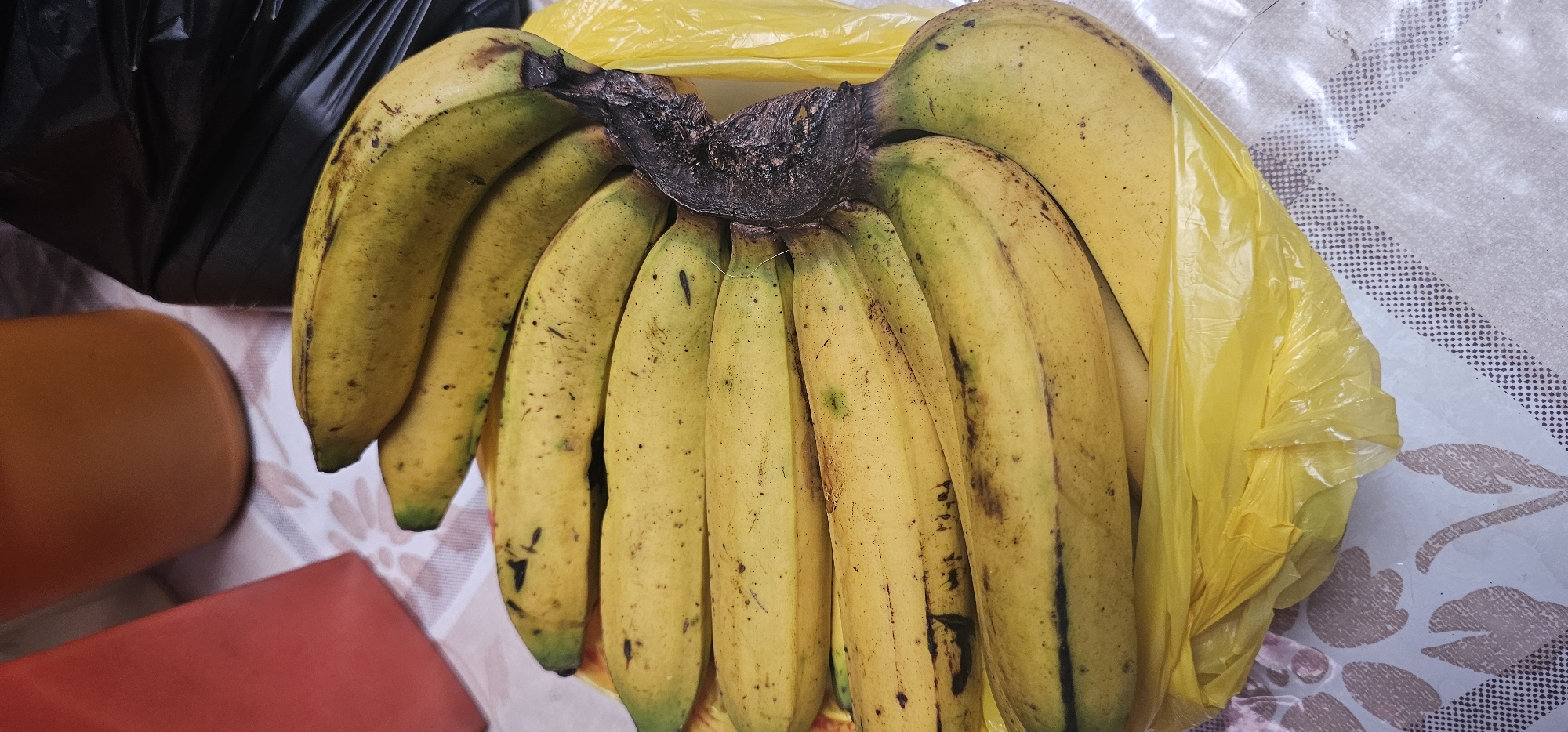 A bunch of ripe yellow bananas is placed in a bright yellow plastic bag. The bananas are slightly speckled with brown spots, indicating their ripeness. The bag is sitting on a patterned surface, possibly a tablecloth, with a white and brown design. In the background, a red object is visible on the left, and a black plastic bag is on the right. The scene appears to be indoors. The image does not provide any specific information about the city of Puncak Jaya, Indonesia.