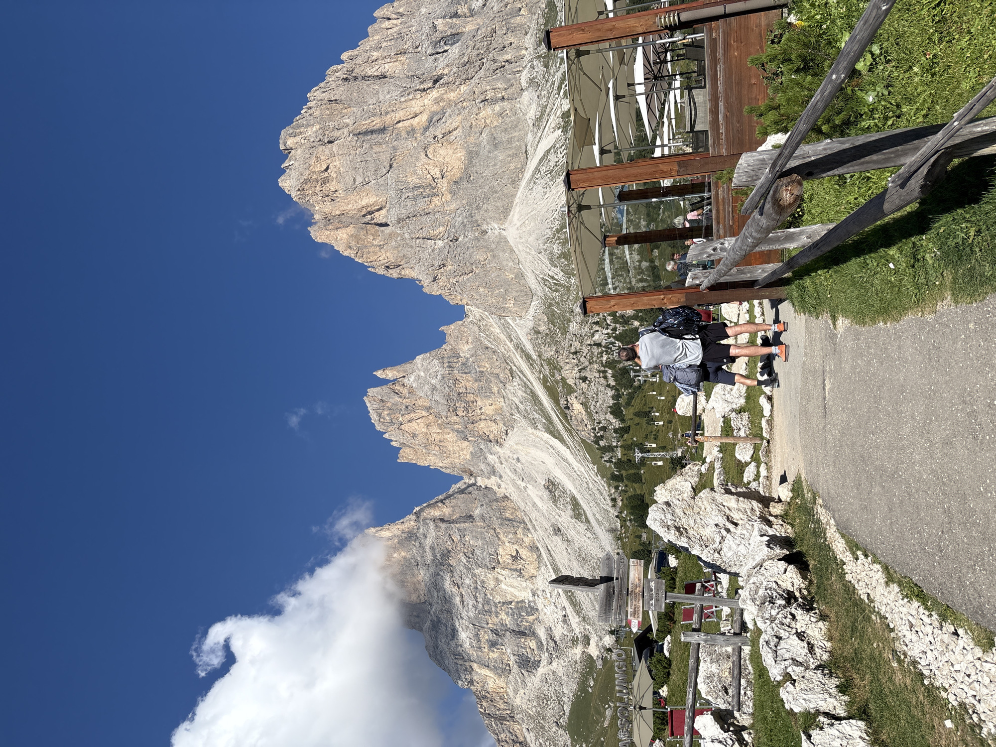 The image shows a view of the Dolomite mountains near Sëlva - Wolkenstein - Selva di Val Gardena, Italy. In the foreground, a paved path leads towards the mountains. To the right of the path is a wooden fence and a wooden structure with several white umbrellas and tables, suggesting an outdoor seating area of a restaurant or cafe. To the left of the path, there is a wooden fence and large rocks. Several people are visible, mostly from behind. One person in the foreground is wearing a grey t-shirt, black shorts, and a backpack, facing away from the camera and towards the mountains. Another person next to them is wearing a dark t-shirt and black shorts. In the midground, more people are seated at tables under the umbrellas. The backdrop is dominated by the imposing rocky peaks of the Dolomites, with some clouds partially obscuring the highest summits. The sky is a clear, bright blue. A ski lift can be seen in the distance on the right side of the image. A wooden signpost with multiple directional arrows is also visible in the midground, closer to the mountains. The overall scene depicts a scenic mountain landscape with tourist amenities.