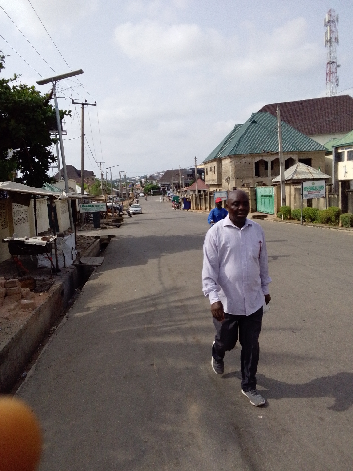 The photograph depicts a street scene in Lokoja, Nigeria. A man in a light-colored long-sleeved shirt and dark trousers is walking toward the camera on an asphalt road. He is wearing gray sneakers. The road is flanked by buildings of varying architectural styles, including a building with a green corrugated roof and another with a brown roof, a sign for "United Evangelical Church" is visible.
On the left side of the road are makeshift stalls and structures. Utility poles with overhead wires line the street. In the distance, another person is riding a motorcycle. A telecommunications tower can be seen in the background. The sky is partly cloudy.