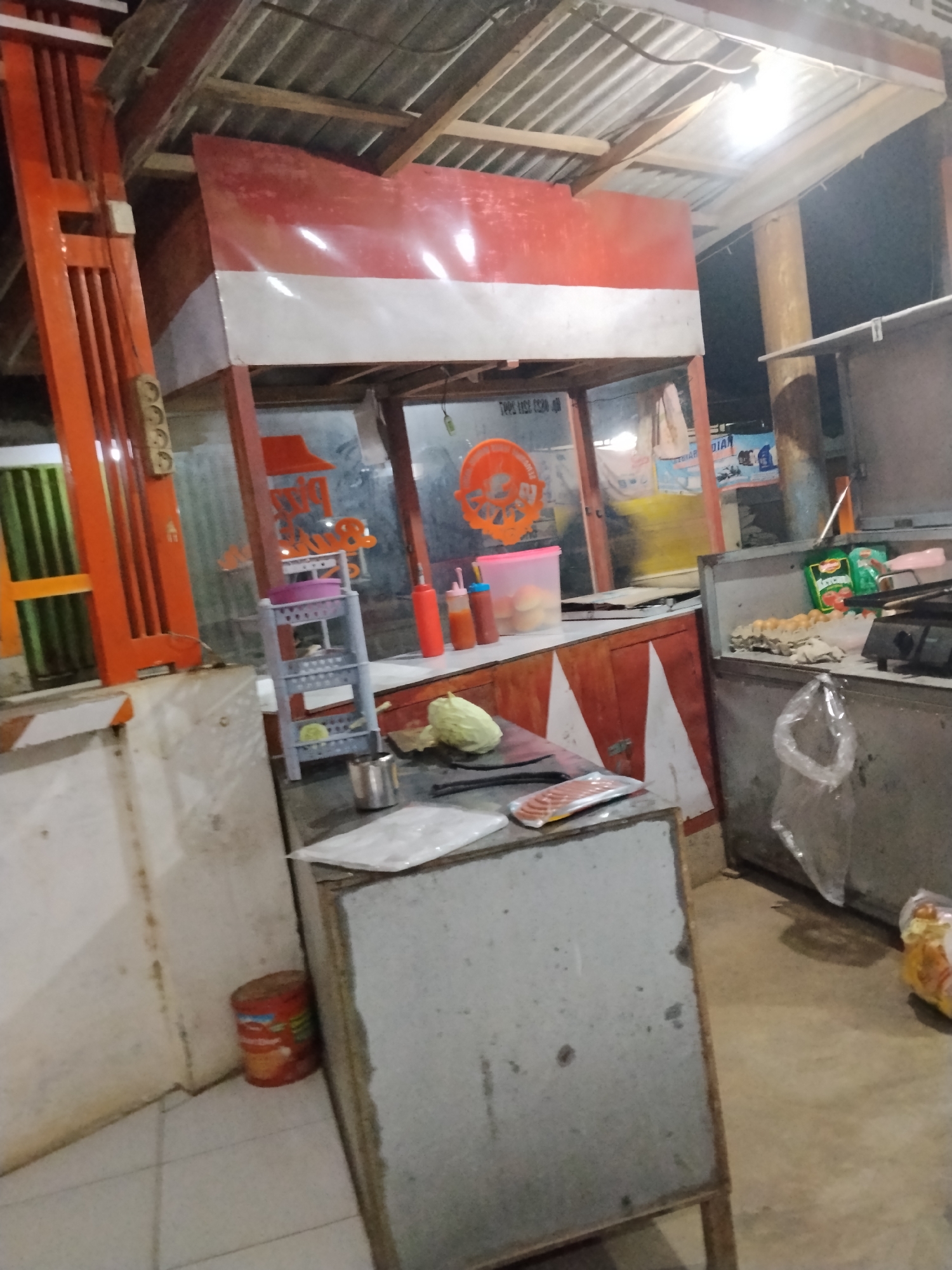 The image shows an outdoor food stall at night. The stall appears to be made of wood and metal, with a red and white awning. There are various food items and cooking equipment visible, including eggs, bottles of sauce, and what looks like raw meat. The stall is lit by artificial lighting, casting shadows on the surrounding area. The ground is paved with tiles, and there are some bags of groceries on the floor. The overall scene suggests a busy food vending operation. The stall's design and the presence of Indonesian text on some signage indicate that this is likely located in Indonesia.