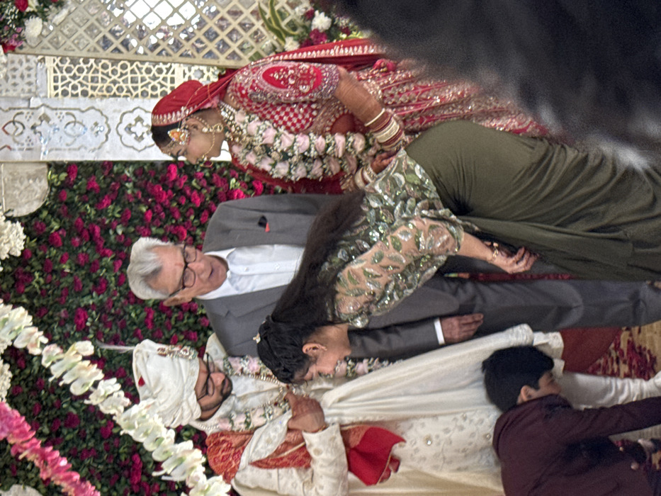 The image captures a vibrant and intimate moment from what appears to be an Indian wedding ceremony, viewed from a slightly elevated perspective. The scene is rich with traditional attire, floral decorations, and family interaction.
In the upper right of the frame is the **bride**, adorned in a traditional red and gold lehenga, intricately embroidered, with a matching red dupatta (veil) draped over her head and shoulders. She wears elaborate gold bridal jewelry, including a maang tikka on her forehead, a nose ring (nath), and earrings. A garland of pink and white flowers is draped around her neck and chest. Her left arm is visible, decorated with several bangles. She is looking downwards or to her left, her expression partially obscured.
In the lower left, seated on the floor, is the **groom**, dressed in a white or off-white sherwani with subtle gold embellishments. He wears a traditional white turban (pagri) adorned with a gold kalgi (turban ornament) and a floral garland similar to the bride's. He has a beard and is wearing glasses. His hands are joined together in front of him, possibly in a gesture of prayer or greeting.
Between the bride and groom, an **older woman** with long dark hair is seen leaning forward. She is wearing a dark olive green outfit, which appears to be a skirt or dress, paired with a long-sleeved top intricately embellished with shimmering, sequin-like patterns. Her right hand is visible near the groom's head, suggesting she is performing a ritual or offering a blessing to him.
Behind the woman in green, to the left, stands an **older man** with short, neatly combed grey hair and glasses. He is dressed in a light grey suit jacket over a white collared shirt. He is looking down towards the groom and the woman, with a gentle smile on his face, indicating warmth and happiness.
A young **child** is partially visible on the far right, seated on the floor next to the groom. The child is wearing a dark maroon or brown shirt and is looking towards the ongoing ceremony. Another person's head is partially obscured by blur on the far right.
The background is elaborately decorated. A prominent wall covered with dense, vibrant red and pink flowers, likely roses, forms a lush floral backdrop behind the standing man and the bride. Above this floral wall, there's an intricate, light-colored lattice-work screen with geometric patterns, possibly made of stone or wood. White floral garlands and green foliage are also visible on the left side of the frame, adding to the festive atmosphere. The floor on the right side of the image features a patterned rug or carpet in shades of red and cream.
The overall scene conveys a sense of celebration, tradition, and family togetherness during a significant life event. The lighting appears soft and even, suggesting an indoor setting or a well-shaded outdoor area. No discernible text is visible in the image.