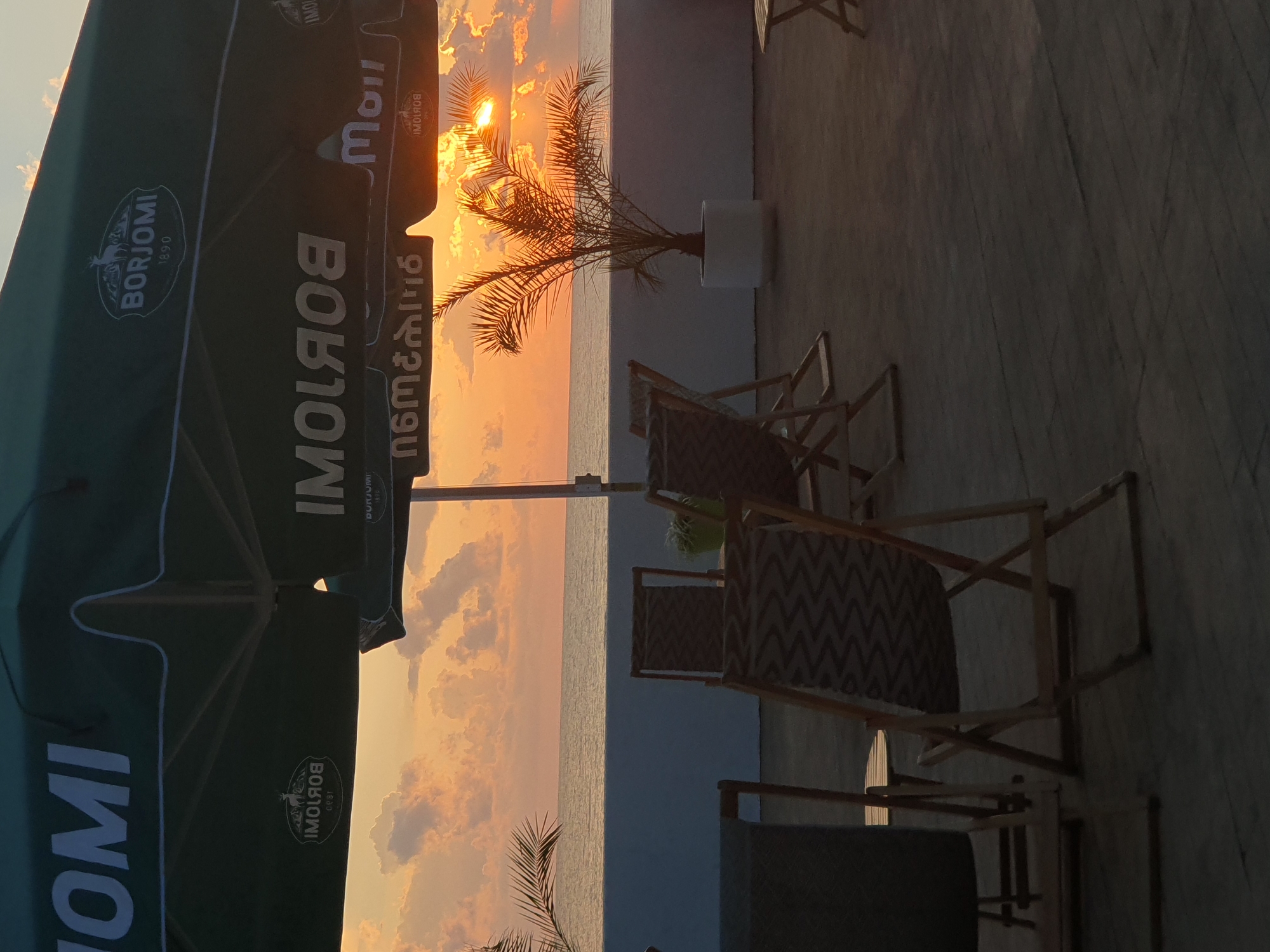This is a photograph taken from a terrace at sunset, likely at an outdoor cafe or restaurant, overlooking the Black Sea. The setting appears to be Batumi, Georgia, suggested by the presence of "BORJOMI" branding on umbrellas, a well-known Georgian mineral water brand, often found in establishments in the region.
In the foreground, several empty beach chairs are arranged on a wooden deck. These chairs have wooden frames and fabric seats with a zig-zag pattern. One chair on the far left is dark and partially obscured. In the mid-ground, a small palm tree in a white planter is situated near the edge of the terrace.
The background features the Black Sea stretching to the horizon, with a clear line dividing the water from the sky. The sky is filled with warm, orange and pink hues characteristic of a sunset, with scattered clouds catching the light. The sun is partially visible, casting a bright glow. The terrace is covered by large, green umbrellas, also bearing the "BORJOMI" logo, which are mostly in shadow. The overall atmosphere is serene and peaceful, with the scene captured during the golden hour. No people are present in the image.