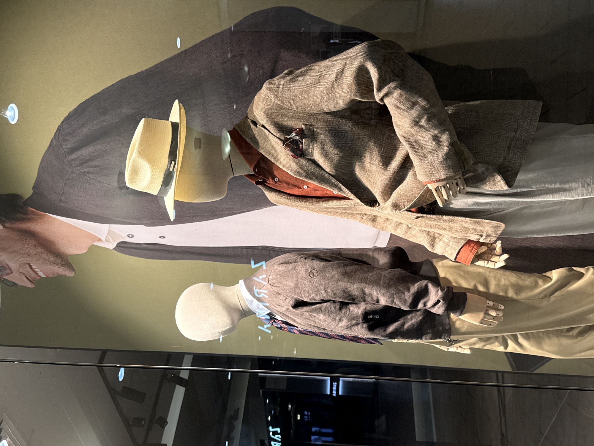 The image depicts a store window display featuring two male mannequins dressed in stylish clothing. The mannequin on the left is wearing a dark brown bomber jacket, a light blue collared shirt, and a striped tie with shades of blue and red. It is also wearing light beige pants. The mannequin on the right is dressed in a light brown patterned blazer, an orange collared shirt, and light gray pants. A straw hat with a black ribbon rests on its head. A pocket square with a patterned design is visible in the blazer's breast pocket. The background of the display features a large photographic print of a man wearing a dark jacket and a white collared shirt, with a portion of his face visible. The overall impression is a sophisticated retail display, likely for men's fashion. The location is identified as Ukromne, Ukraine, suggesting this is a storefront in that city.
