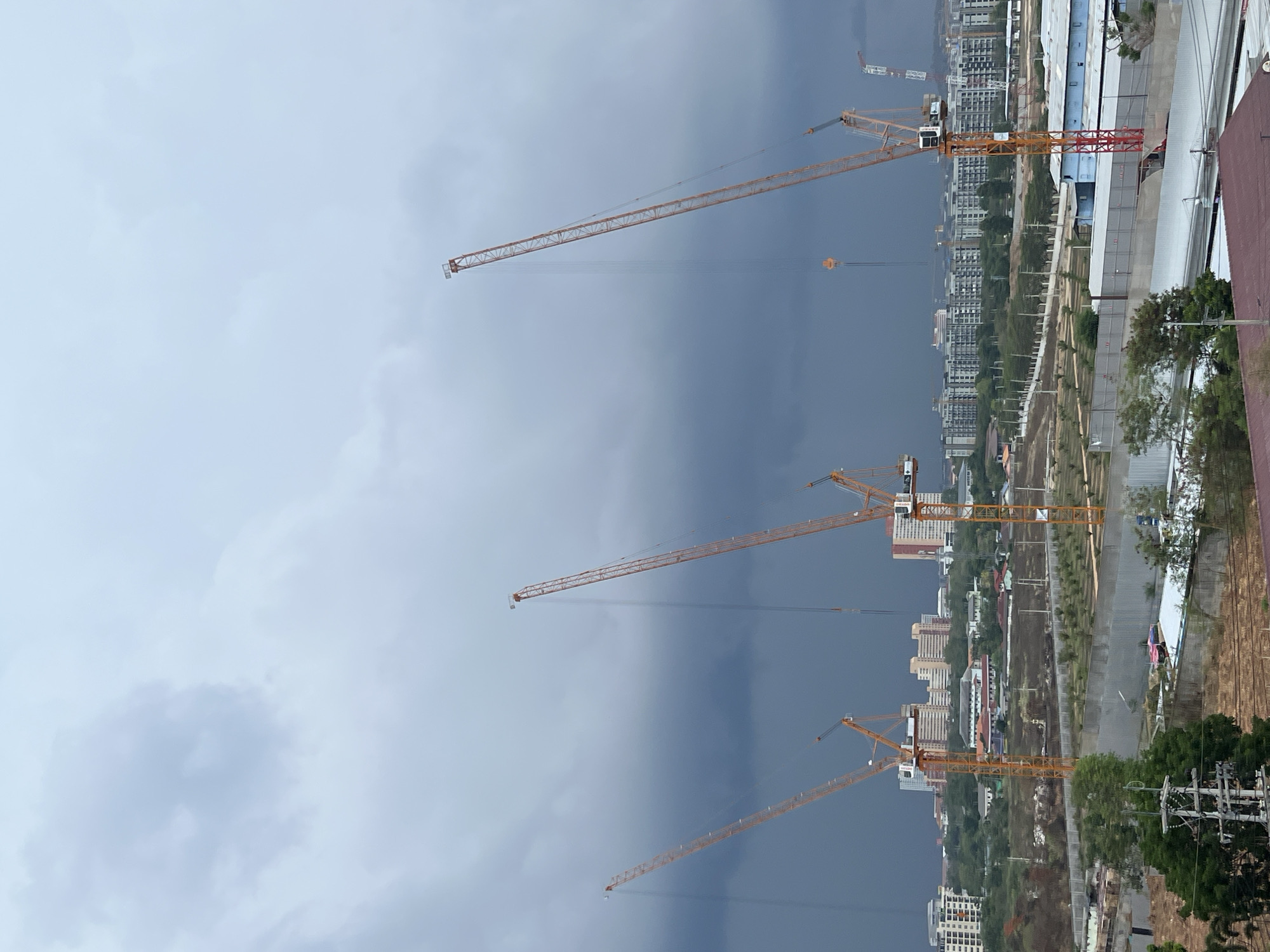 The image captures a cityscape in Pattaya City, Thailand, dominated by several construction cranes against a backdrop of a stormy sky. The cranes, identifiable by their orange lattice structures, suggest ongoing construction projects. The city's landscape includes numerous high-rise buildings and greenery, indicating a mix of urban and natural environments. The overcast sky and dark clouds imply imminent rain or a recent storm.