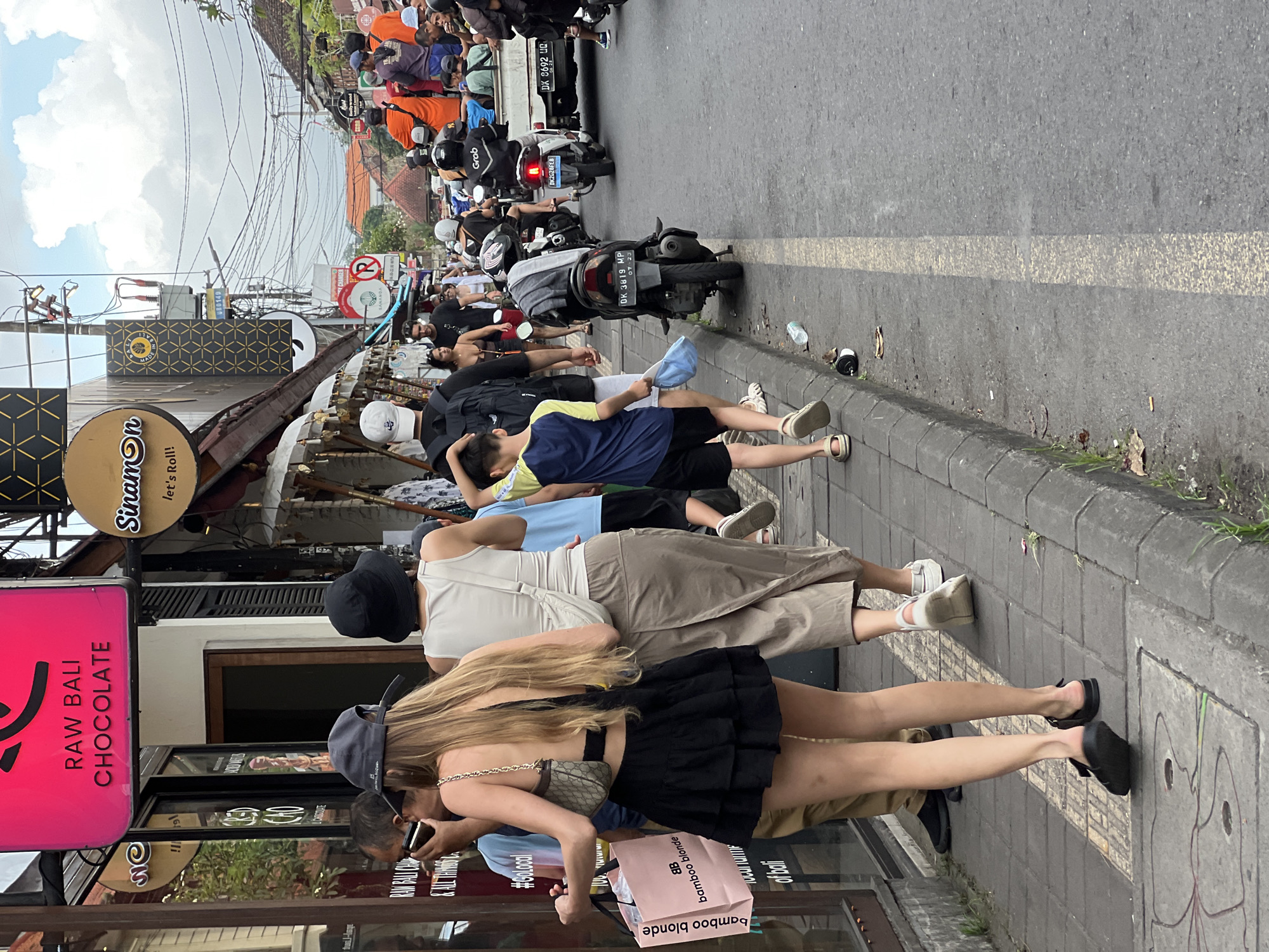 The image is a street scene in the Ubud District of Indonesia, specifically on a narrow road packed with motorbikes and pedestrians. On the left side, storefronts are visible, including "Raw Bali Chocolate" and "Sinamon let's Roll!" A line of people is standing or squatting in front of the shops, some looking at their phones. They include a woman with long blond hair holding a "bamboo blonde" shopping bag. A child in a blue shirt stands among them. The street is full of motorbikes and pedestrians, indicating traffic congestion. Overhead, there are many tangled power lines.