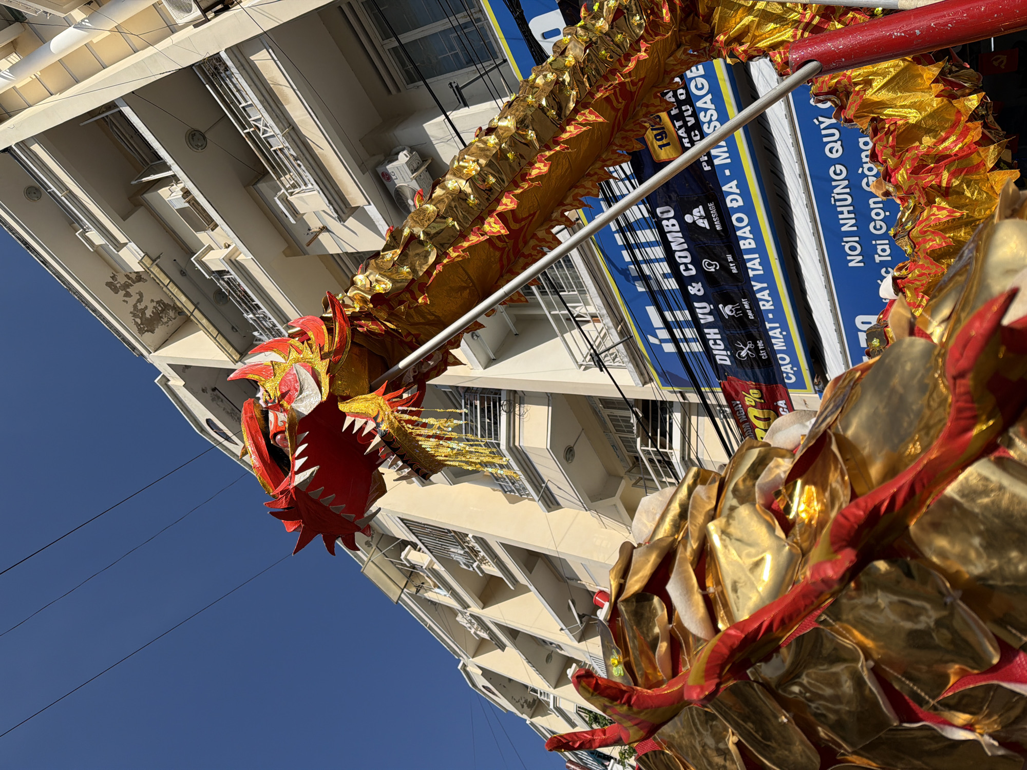 In Nha Trang Ward, Vietnam, a close-up, low-angle shot captures a vibrant, golden dragon used in a street celebration. The dragon, primarily red and gold, is the central focus, its scales shimmering under the sunlight. The dragon's head, with its sharp teeth and ornate red and gold detailing, is in the upper left of the frame. The long, segmented body of the dragon extends across the majority of the image, with its gold scales detailed with red accents.
In the background, a pale yellow, multi-story building dominates the scene. The building features numerous balconies with metal railings and several windows. The architecture suggests an urban environment. Several electrical wires are visible against the clear, bright blue sky, crossing from the upper left to the upper right of the image.
Below the building, a bright blue banner with white text is displayed. The text appears to be in Vietnamese and includes phrases like "DICH VU & COMBO," "MASSAGE," and "CAO MAT - RAY TAI BAO DA." Another blue banner with white text below it reads "NOI NHUNG QUY" and "Tai Gon Gòng." These banners indicate commercial establishments, possibly a spa or salon. The presence of the dragon and banners suggests a festive occasion, such as a holiday parade or cultural event. The lighting indicates it is daytime, with strong sunlight casting shadows. There are no visible people in the frame.