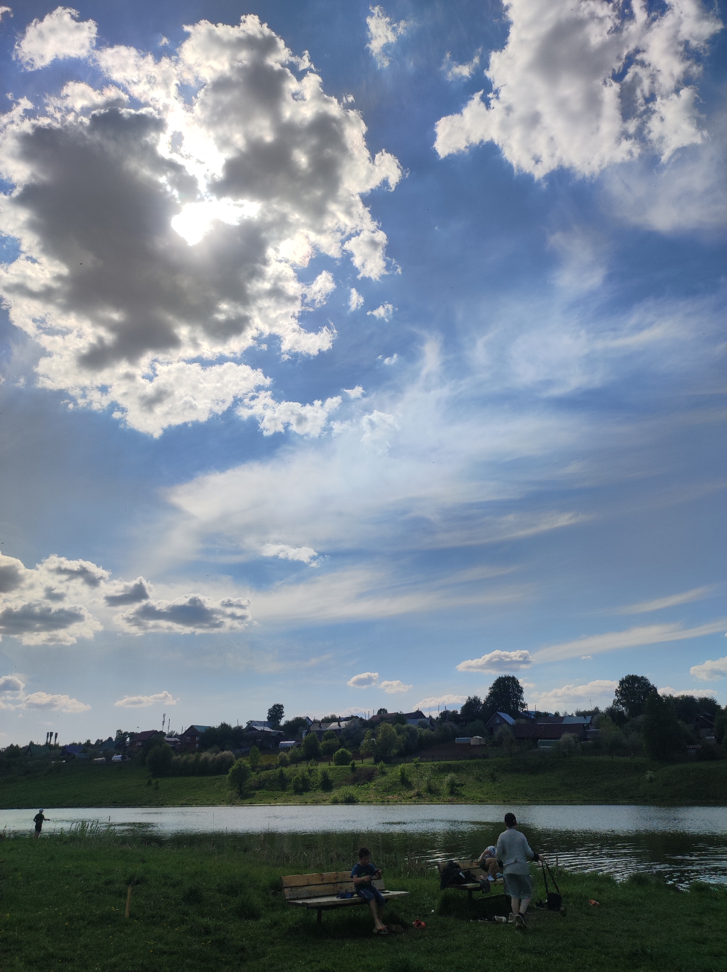 The image depicts a riverside scene in the city of Фоки, Russia. In the foreground, a grassy bank leads to a body of water. Two benches are positioned on the grass; one is occupied by a person seated alone, and the other by two other people. A man is standing, appearing to be fishing or preparing to fish. A person stands near the water on the left side of the image. The middle ground features the body of water. Beyond the river, a hill rises, topped with a row of houses and vegetation, suggesting a village or residential area. The background is dominated by a bright sky with cumulus clouds and cirrus clouds, with the sun partially obscured behind the clouds.