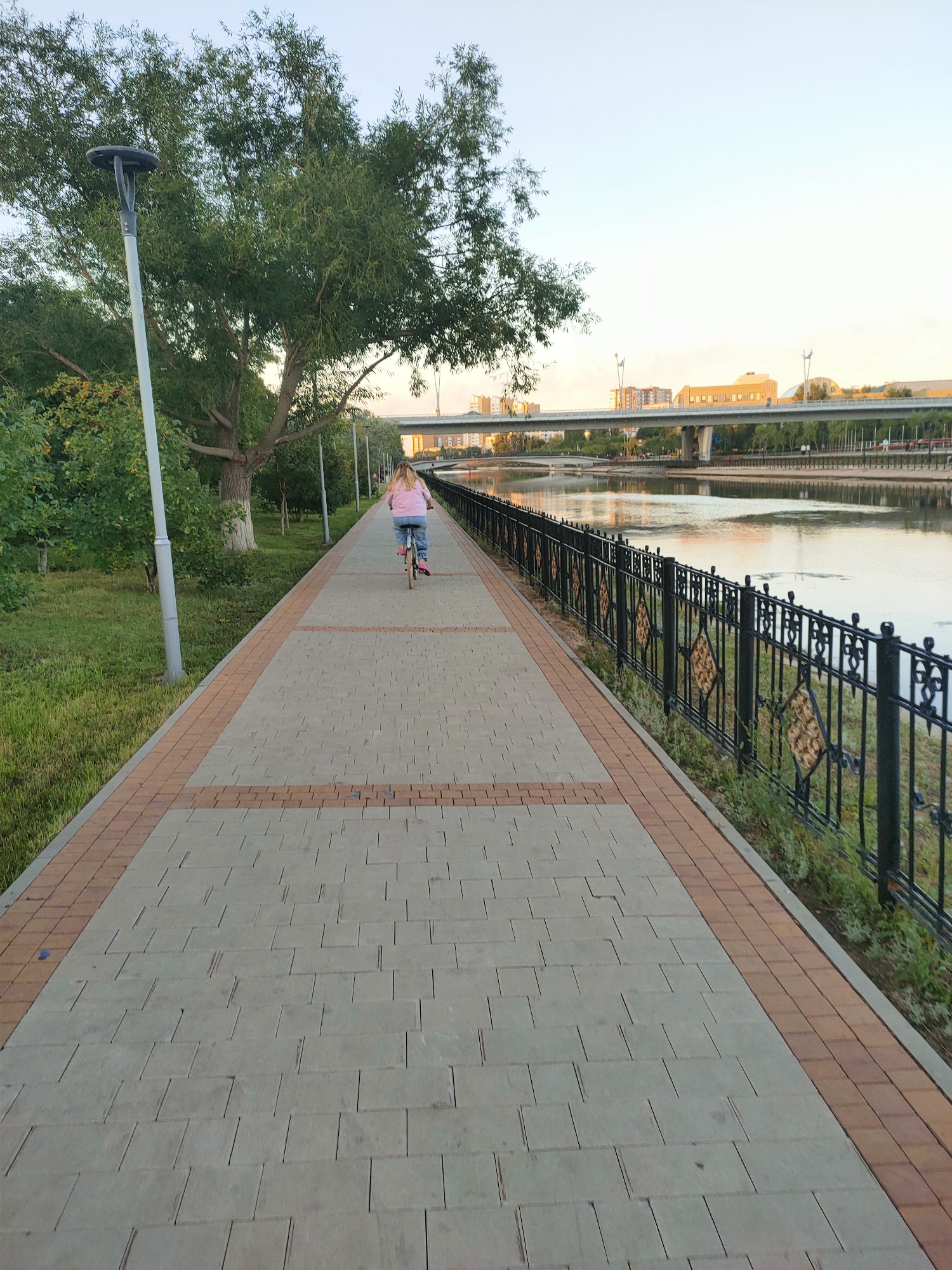The image shows a view of a pedestrian and bicycle path along the Ishim River in Astana, Kazakhstan. A person is riding a bicycle on the path, facing away from the camera. The path is paved with grey and brown rectangular tiles. To the left of the path is a grassy area with trees and a lamppost. To the right of the path is a black metal fence with decorative gold diamond shapes. The Ishim River is visible to the right of the fence, with a bridge and buildings in the background. The sky is clear and light blue.