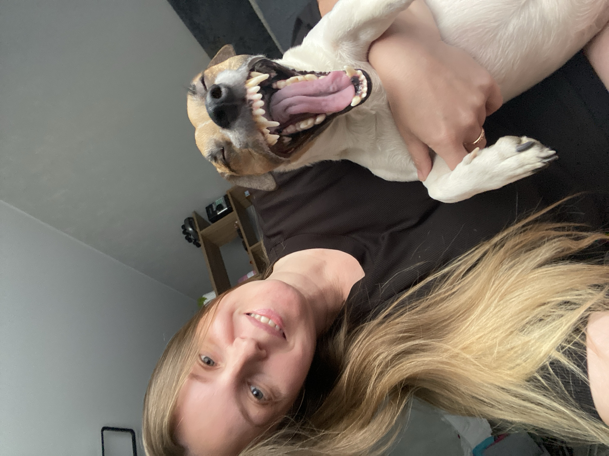 A selfie photo shows a woman with long blonde hair, wearing a black shirt, holding a small dog. The dog is held upside down and is showing its teeth and pink tongue. The woman is smiling and looking at the camera. In the background is a gray wall and a brown shelf unit with various items on it. The location is indoors. The city of Noviy, Russia is not discernable from the image.
