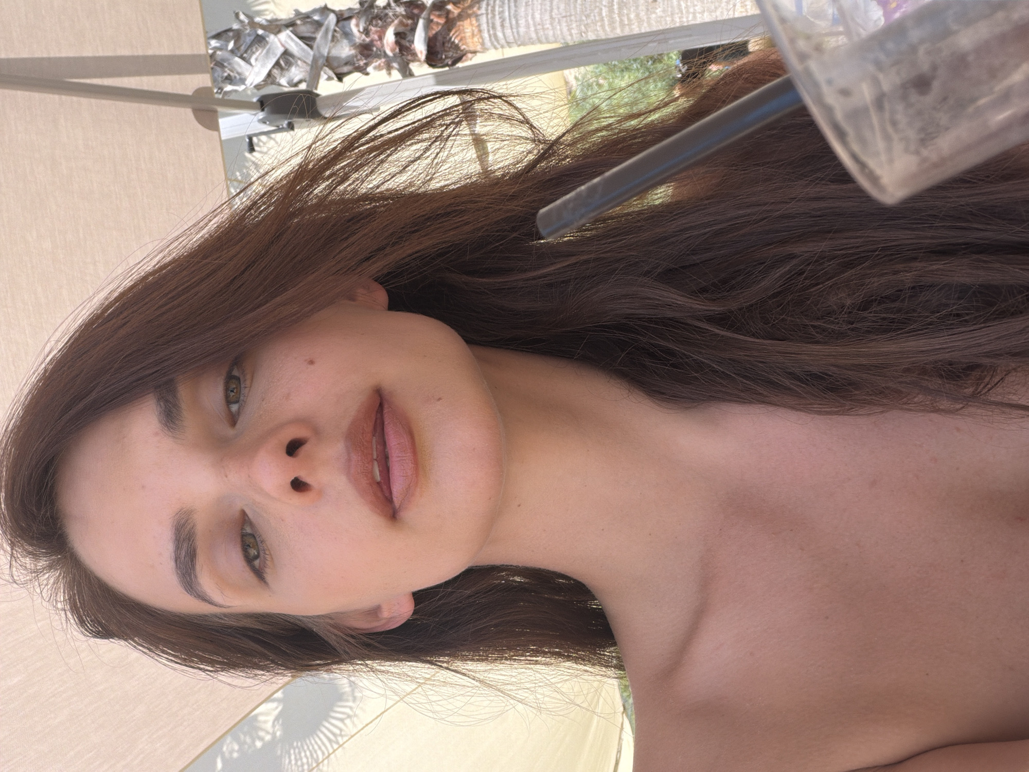 A young woman with long, dark brown hair is shown in a close-up, outdoor selfie. She is looking up towards the camera with a neutral expression. Her hair is partially obscuring her face. She is wearing a strapless top, revealing her shoulders and upper chest. In the foreground, a clear plastic cup with a dark straw is visible on the right side of the image. The background suggests an outdoor setting with a palm tree and what appears to be a large, light-colored umbrella providing shade. The scene is brightly lit, indicating it is daytime. The location appears to be Fethiye, Turkey, possibly at a beachside resort or cafe, given the presence of the palm tree and umbrella.