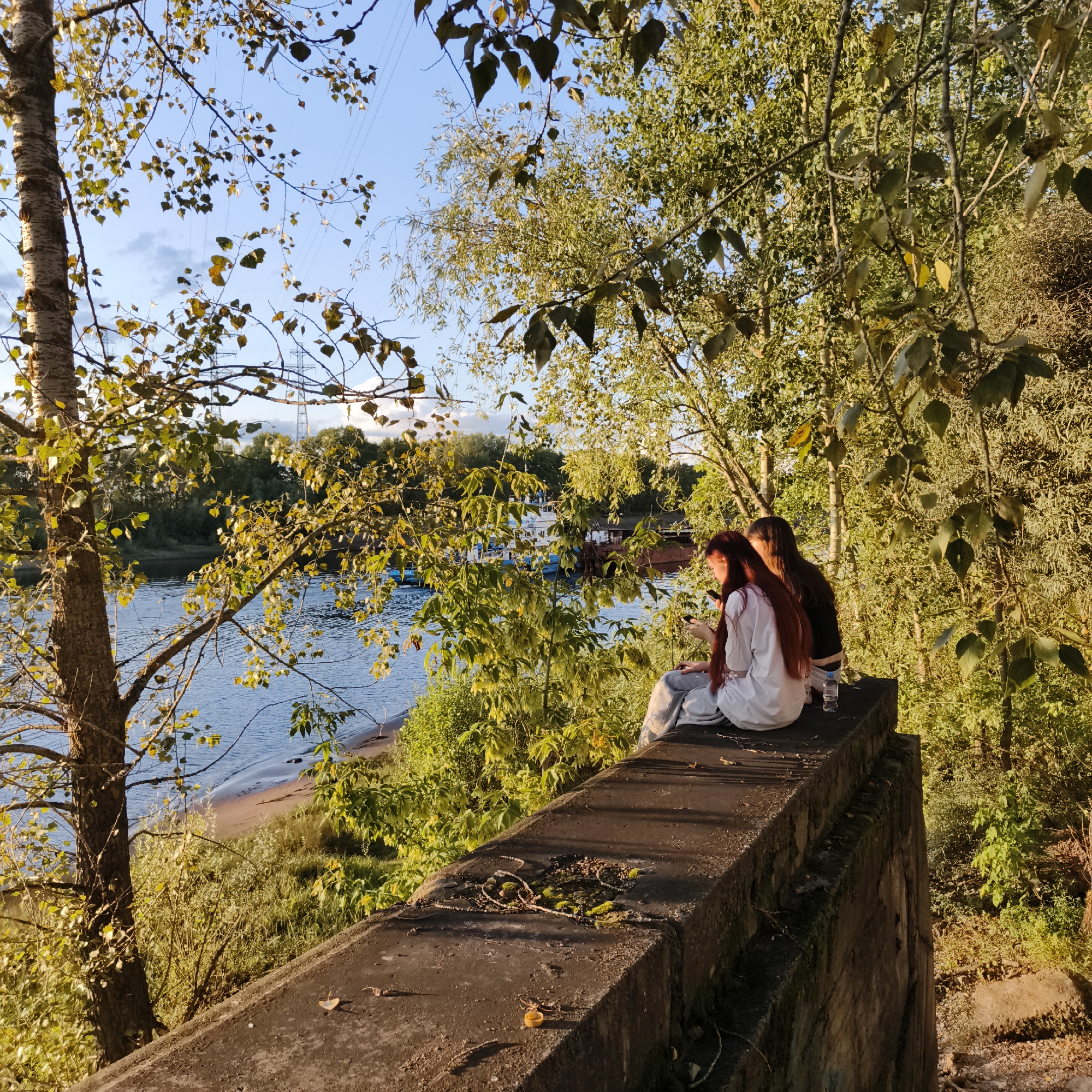 In Chaikovsky, Russia, two young women are seated on a concrete structure overlooking the Kama River during what appears to be late afternoon or early evening. The woman on the left has long red hair and is wearing a white t-shirt and grey sweatpants, holding a smartphone in her hands. The woman on the right has dark hair and is wearing a black t-shirt and dark pants, also looking at her phone. A plastic water bottle is placed on the concrete beside them. The surrounding area is lush with green trees, some of which have autumnal yellowing leaves, indicating a transition into autumn. The river is a calm blue, with a sandy bank visible on the opposite side. In the distance, several boats are moored on the water. The overall scene suggests a peaceful, casual moment of relaxation by the river, with power lines visible in the sky, hinting at urban proximity.