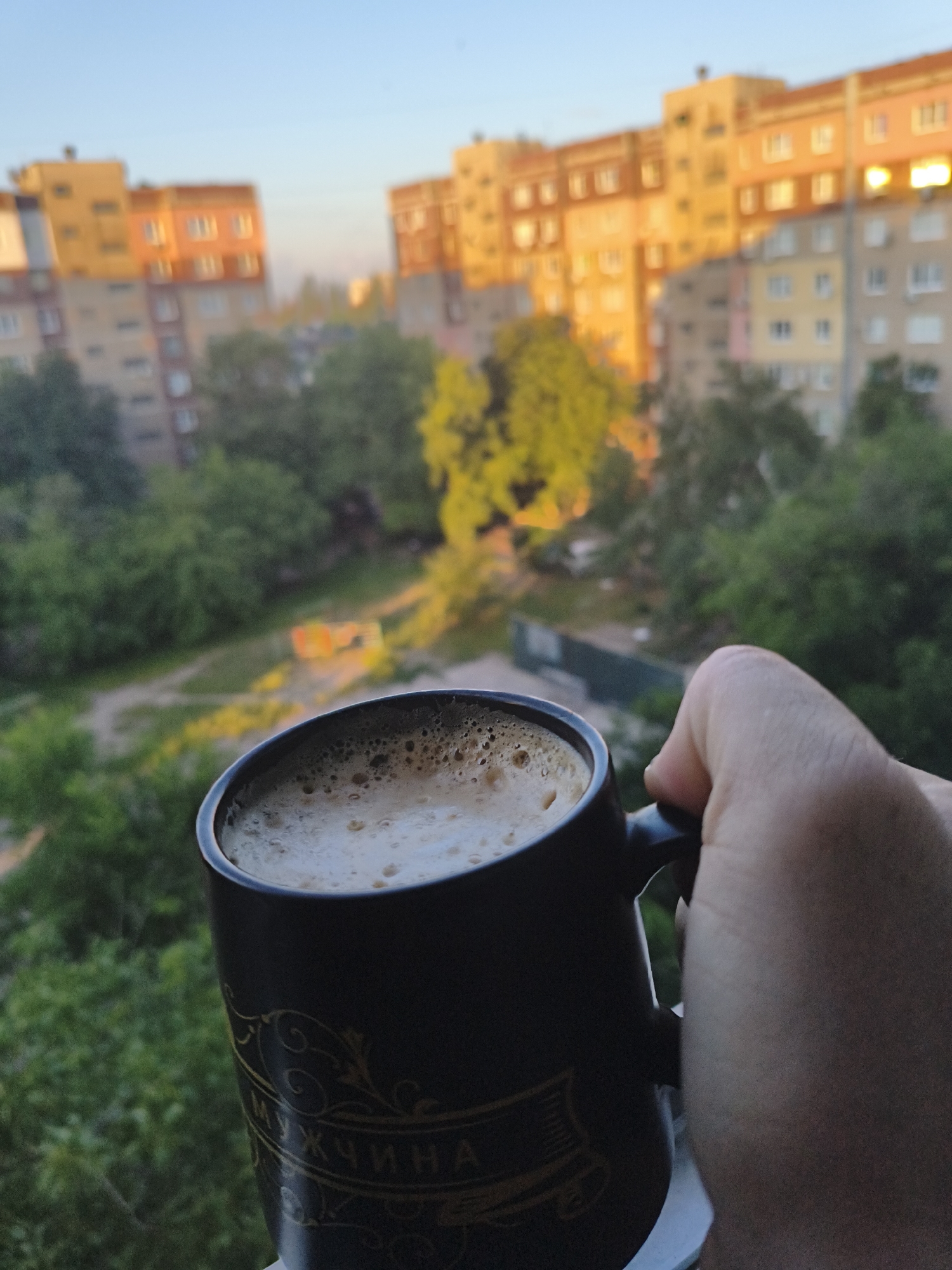 A hand holds a dark mug of coffee with a frothy top. The mug has decorative gold lettering on it. In the background, apartment buildings are visible, bathed in the warm light of what appears to be sunset. Green trees and foliage surround the buildings. The scene is set against a clear blue sky.