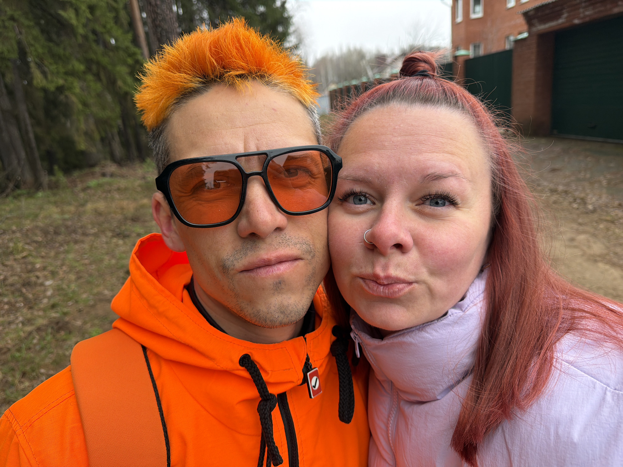 A close-up selfie captures a man and a woman posing outdoors in Chaikovsky, Russia, on what appears to be an overcast day.
The man, positioned on the left, has striking bright orange, spiky hair and is wearing dark-framed sunglasses with orange-tinted lenses. He has light stubble and is dressed in a vibrant orange hooded jacket, with a matching orange backpack strap visible over his left shoulder. A small red tag with a white checkmark symbol is attached to his jacket's zipper pull. He looks directly at the camera with a neutral to slightly serious expression.
The woman, on the right, has reddish-brown hair styled in a high bun. She has light blue eyes and a silver ring piercing in her left nostril. She is wearing a light pink or lavender puffy jacket and is captured mid-pucker, suggesting a playful expression for the photo.
The background shows a dense cluster of green trees on the left side, indicating a forested or natural area. On the right, behind the woman, there is a red brick building with a dark green gate or garage door, suggesting a residential or industrial structure. The sky overhead is cloudy.