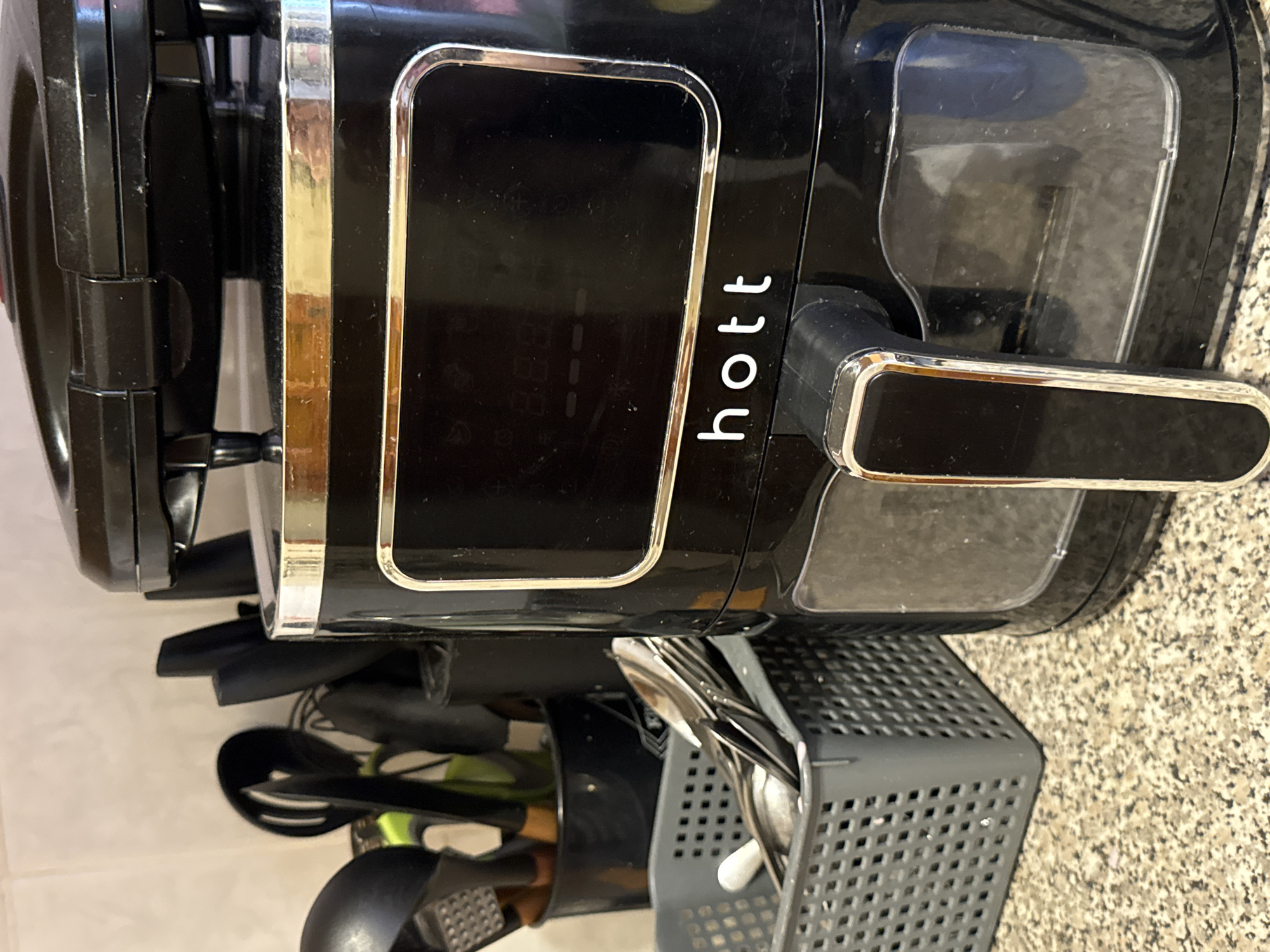 The image shows a black air fryer on a speckled countertop. To the left of the air fryer is a gray basket filled with silverware. Behind the basket, a black container holds various kitchen utensils, including a slotted spoon, a whisk, and a grater. The background consists of a beige tiled wall. The air fryer has a digital display and a silver trim around it. The word "hott" is visible in white lettering on the front of the air fryer. The overall setting appears to be a kitchen. There are no people or animals present in the image. The city of Яромінскі сельскі Савет, Belarus is not visible in the image.