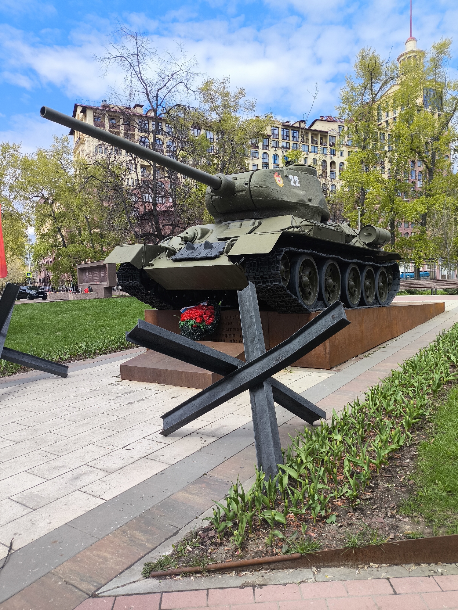The image shows a T-34 tank monument in a park-like setting in Moscow, Russia. The tank, painted in olive green, is mounted on a rust-colored plinth. A red star emblem and the number "22" are painted on the turret. Anti-tank obstacles, known as Czech hedgehogs, are placed in front of the monument. A wreath of red flowers sits below the tank. The surrounding area includes sidewalks paved with stone tiles, flowerbeds with sprouting tulips, green lawns, and trees with budding leaves. In the background, multi-story residential buildings are visible under a blue sky with scattered clouds.