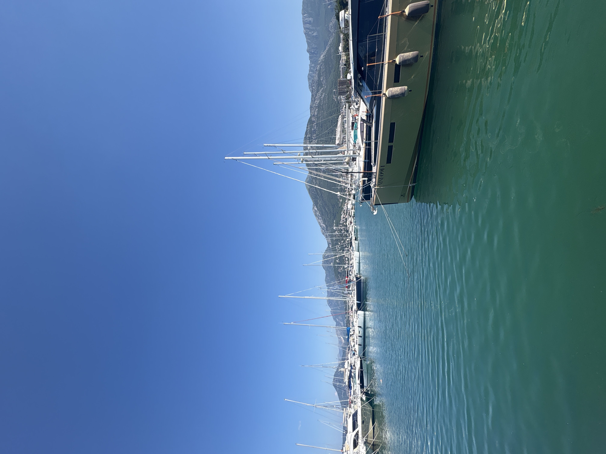 This is a photograph of a marina in Bar, Montenegro. The image is taken from the water's surface, looking towards a line of sailboats and a larger motor yacht docked in the foreground. The water is a clear, turquoise color and shows gentle ripples. In the background, a range of green, mountainous terrain is visible under a bright blue sky. The boats are all white with tall masts, except for the motor yacht in the foreground, which is a light olive green with tinted windows. The motor yacht has the registration number "MNE0911" visible on its side. There are no people visible in the image. The scene suggests a calm and sunny day at the marina.