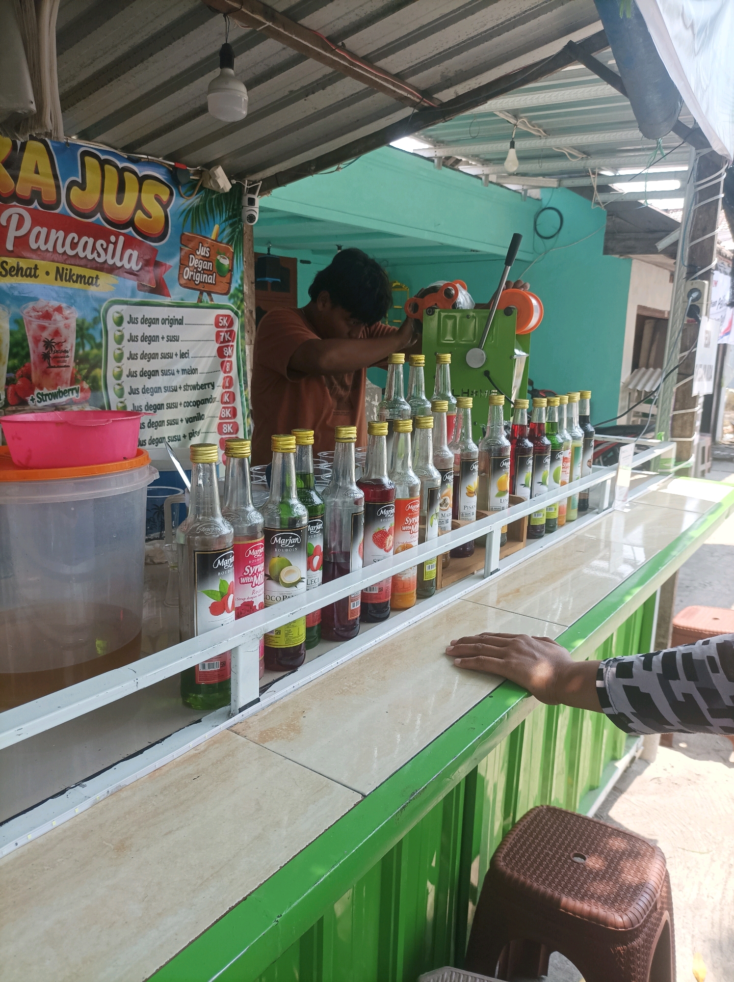 This is a medium shot of an outdoor juice stand located on a street in Sumuragung, Indonesia. A man, possibly the vendor, is seen leaning over a green ice-shaving machine. A large plastic container filled with liquid sits on the left side of the counter, topped with a pink bowl. A row of colorful syrup bottles, mostly from the "Marjan" brand, are neatly arranged on the counter. The juice stand has a striped awning and a banner advertising "Jus Pancasila." The background shows a turquoise wall and a glimpse of a residential building. A customer's hand rests on the counter in the foreground, and a brown plastic stool is visible near the bottom right. The scene depicts a typical street-side business operation in Indonesia.