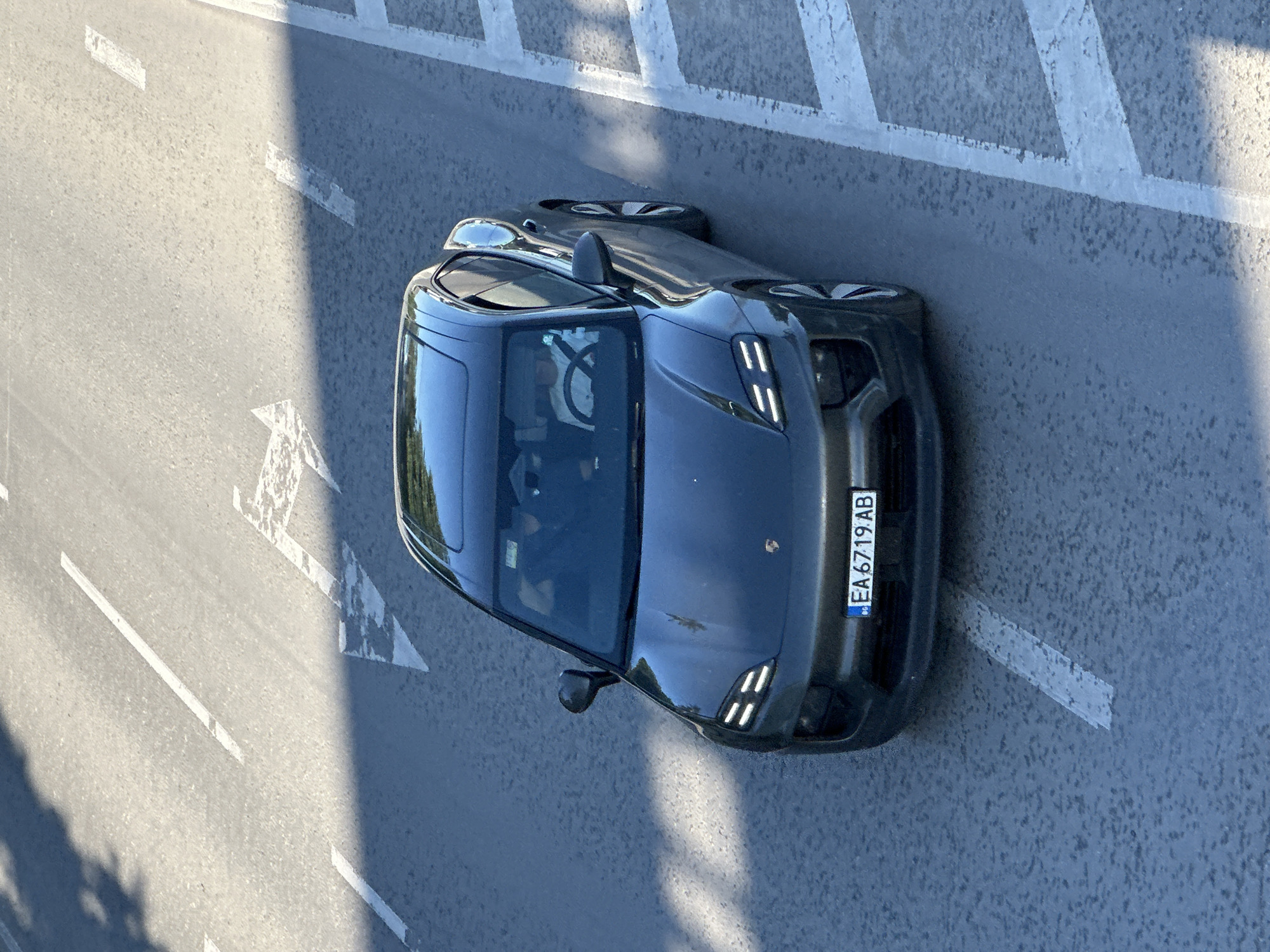 The image shows a dark-colored car, possibly a Porsche Taycan based on its distinctive headlight design, parked or driving on a road. The license plate is visible, reading "EA 6719 AB." The road has painted lane markings and possibly crosswalk indicators. Based on the plate code, the car is located in Bulgaria, but Veliko Tarnovo cannot be confirmed from this image alone. There are people inside the car, but details are difficult to discern. The environment appears to be a city street.