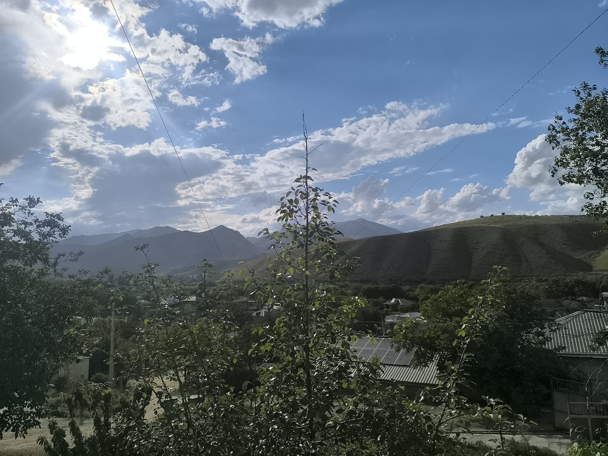 This image captures a picturesque, elevated view of a rural mountainous landscape in Areni, Armenia, under a dynamic sky. The scene is dominated by natural elements, with hints of human settlement.
In the foreground, lush green foliage from several deciduous trees occupies a significant portion of the frame. Their vibrant leaves are well-lit, some reflecting the bright sunlight. A prominent, slender tree with sparse branches and green leaves stands centrally, reaching towards the sky.
The mid-ground reveals a collection of houses or structures nestled within the landscape. Several homes with distinctive corrugated metal roofs are visible, and at least two are equipped with solar panels, suggesting a blend of traditional architecture and modern energy solutions. Hills and valleys covered in green vegetation frame these settlements.
In the background, a majestic range of mountains and rolling hills stretches across the horizon. Some hills are richly green, while more distant peaks appear hazy and bluish-grey, indicating depth and scale.
The sky above is a striking combination of clear blue patches and dramatic white and grey clouds. The sun, partially obscured by clouds in the upper left, emits bright rays and a noticeable lens flare, suggesting a late morning or early afternoon time. Utility lines are visible, traversing the sky diagonally across the frame.
The overall impression is one of serene natural beauty in a rural Armenian setting. No people or visible text are present.