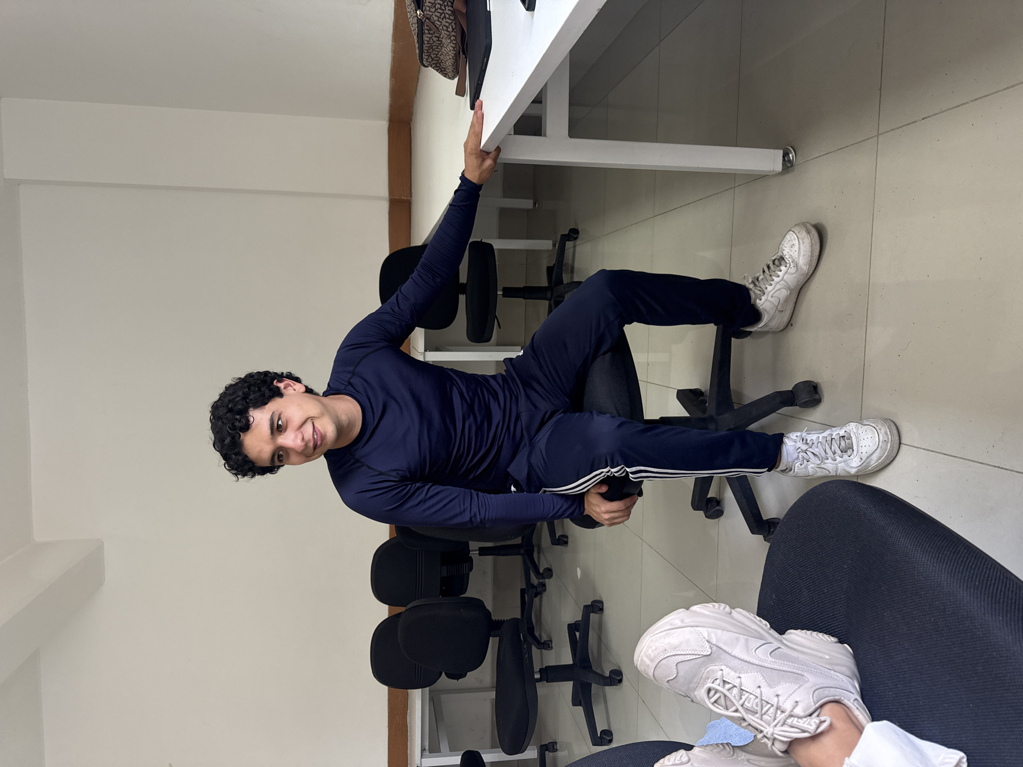 The image shows a young man with dark curly hair, wearing a navy blue long-sleeved shirt and pants with a white stripe down the side, sitting on an office chair in what appears to be an office setting. He is smiling at the camera and holding onto the edge of a white desk with his right hand. His feet are clad in white sneakers.
Several other black office chairs are visible behind him. The floor is tiled in a light color. The location is presumed to be San Andrés Cholula, Mexico. A glimpse of another person's lap and feet, clad in white sneakers, can be seen at the bottom right corner of the frame.
