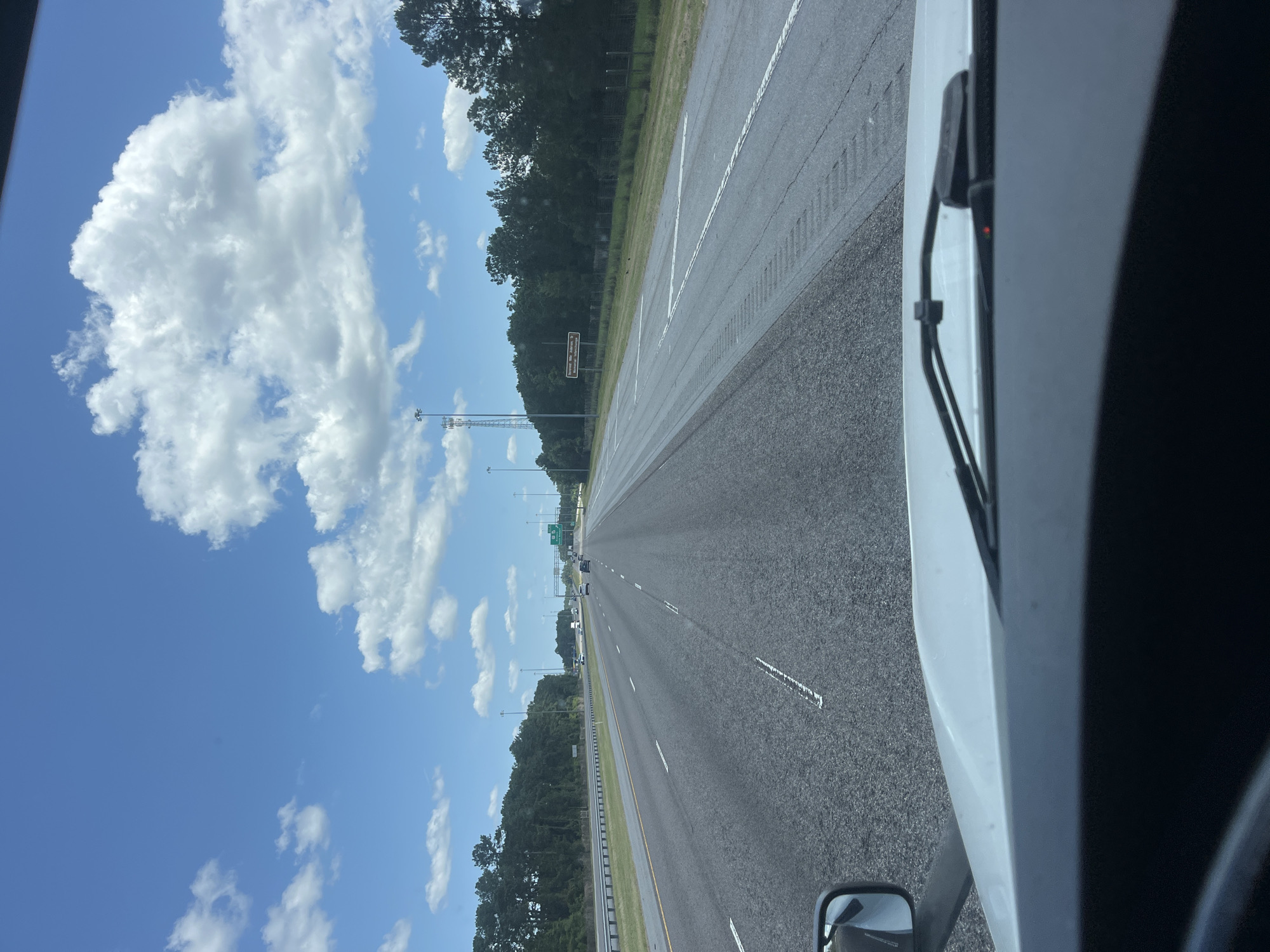 This is a POV shot from the interior of a vehicle, looking out at a highway in Shellman Bluff, Georgia, United States. The road is asphalt with white lane markers and appears to be a multi-lane highway with cars driving in the distance. To the left of the road is a grassy median and then a line of trees. To the right of the road, there is also a grassy area with trees and a sign that reads "Evergreen Villa Anderson St Evergreen Interchange". The sky is blue with scattered white clouds, and there are several tall cell towers visible in the distance. The front windshield and dashboard of the vehicle are visible in the foreground.