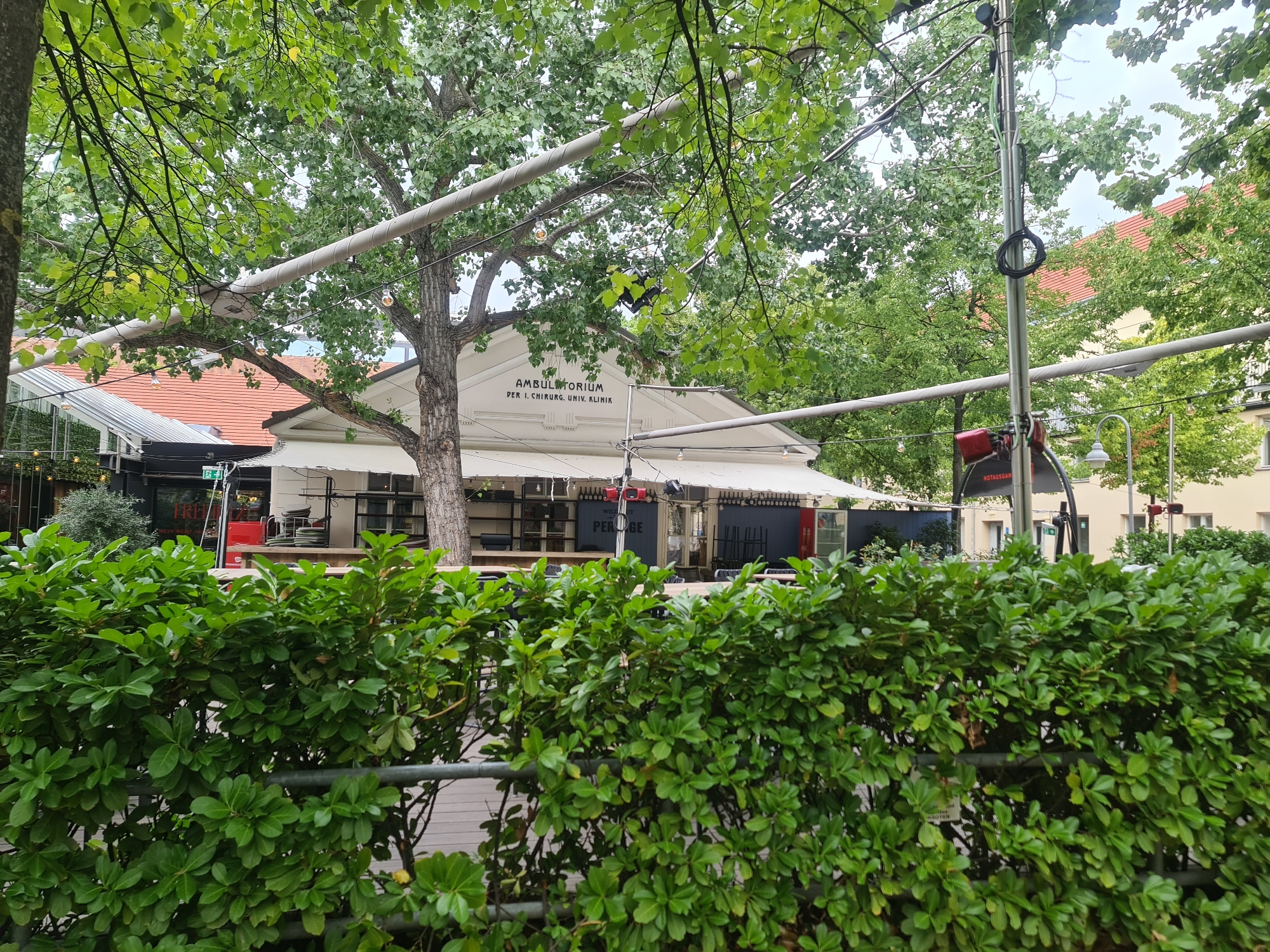 The image shows the outdoor seating area of a restaurant or cafe. In the foreground, there is a dense green hedge. Behind the hedge, there are tables and chairs set up on a wooden deck. A white awning covers part of the seating area. Above the awning, there is a building with the words "AMBULATORIUM PER I. CHIRURG. UNIV. KLINIK" written on it. The building appears to be part of a university clinic. The overall environment suggests a pleasant outdoor dining or gathering space in a city, likely during daylight hours, with trees providing shade. There are no people visible in the image. Based on the architectural style and the text on the building, this location is likely the Ambulatorium of the First Surgical Department of the University Clinic in Vienna, Austria.