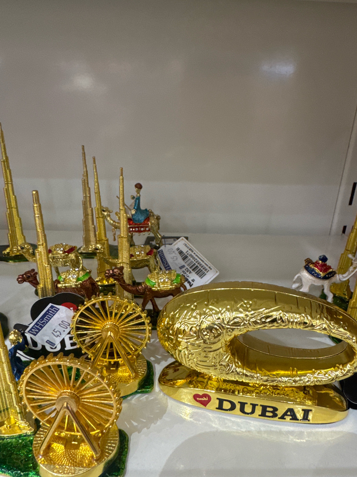 The image displays a collection of gold-colored miniature souvenirs, likely from Dubai, United Arab Emirates, arranged on a white shelf. Prominently featured are replicas of the Burj Khalifa, camels with riders, and Ferris wheels. A large, oval-shaped golden object with Arabic script and the word "DUBAI" inscribed in black lettering on a red heart shape is positioned in the foreground. One of the souvenirs has a white price tag attached, showing "WHSmith 45.00". The background is a plain, light-colored wall with some diffused lighting. The overall scene suggests a retail display of tourist memorabilia. There are no people visible in the image.