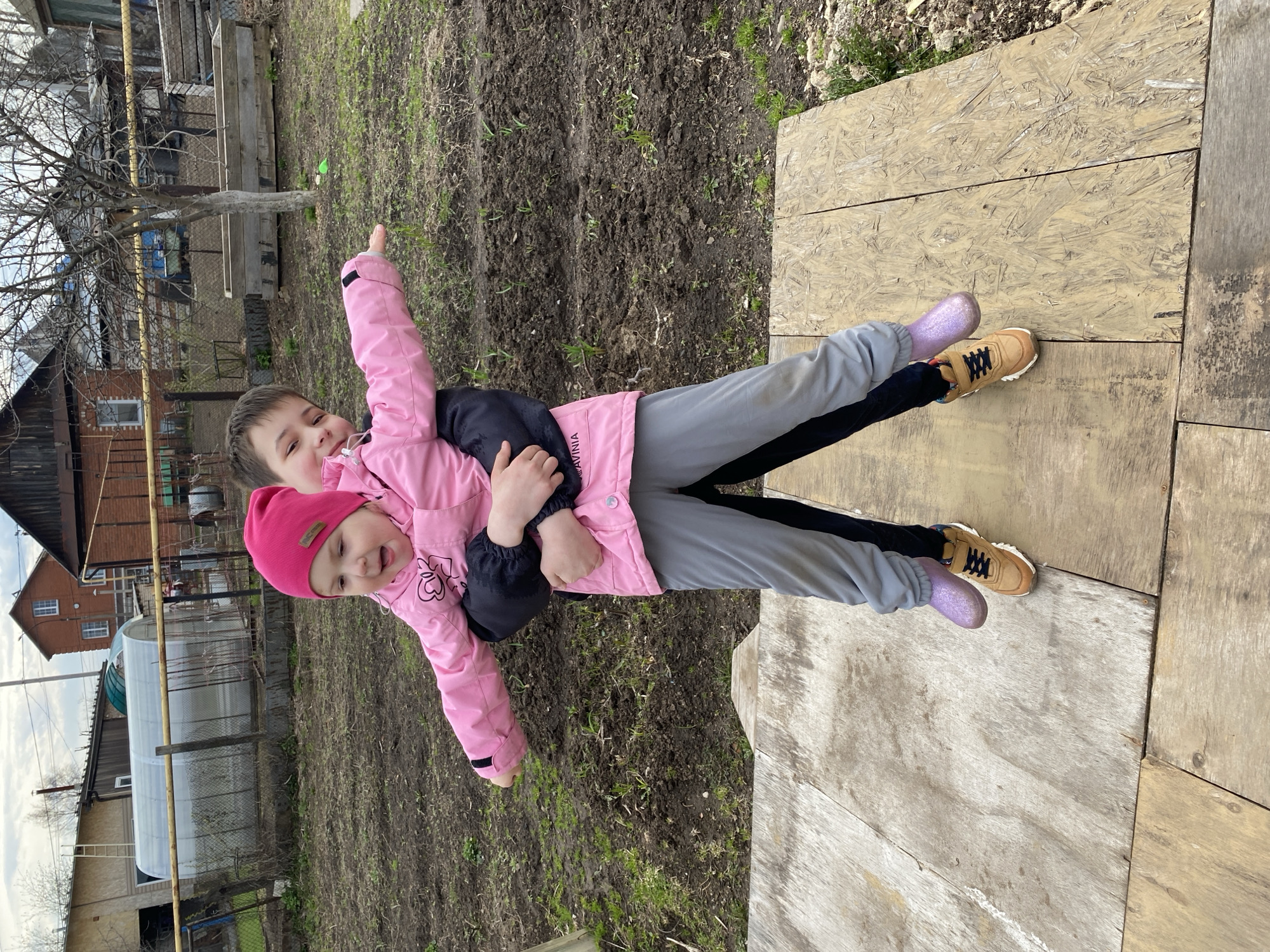 This image depicts two children interacting playfully outdoors in a rural or semi-rural setting, likely in Chaikovsky, Russia. The photograph appears to be taken during the daytime under an overcast sky, with the presence of bare trees suggesting early spring or late autumn.
The older child, with dark hair, is wearing a pink jacket with black sleeves, grey sweatpants with black stripes, and light brown sneakers with black laces. This child is holding the younger child, who is dressed in a bright pink hat, a pink jacket with heart outlines on the chest, and shiny purple glitter boots. Both children are smiling, conveying a sense of happiness and enjoyment. The older child extends their right arm, pointing towards something out of view.
They are standing on a light-colored wooden platform or pathway made of large plywood sheets. Adjacent to this platform is an area of dark, tilled earth with scattered green sprouts. The background reveals various structures common to a village or countryside, including a brick house with white-framed windows, a white greenhouse, and other wooden buildings and fences. Bare trees without leaves are visible in the distance, contributing to the cool, outdoor ambiance.
Visible text includes "AVINIA" printed on the left sleeve of the older child's pink jacket. The overall scene captures a moment of joy and interaction between the two children in their outdoor environment.