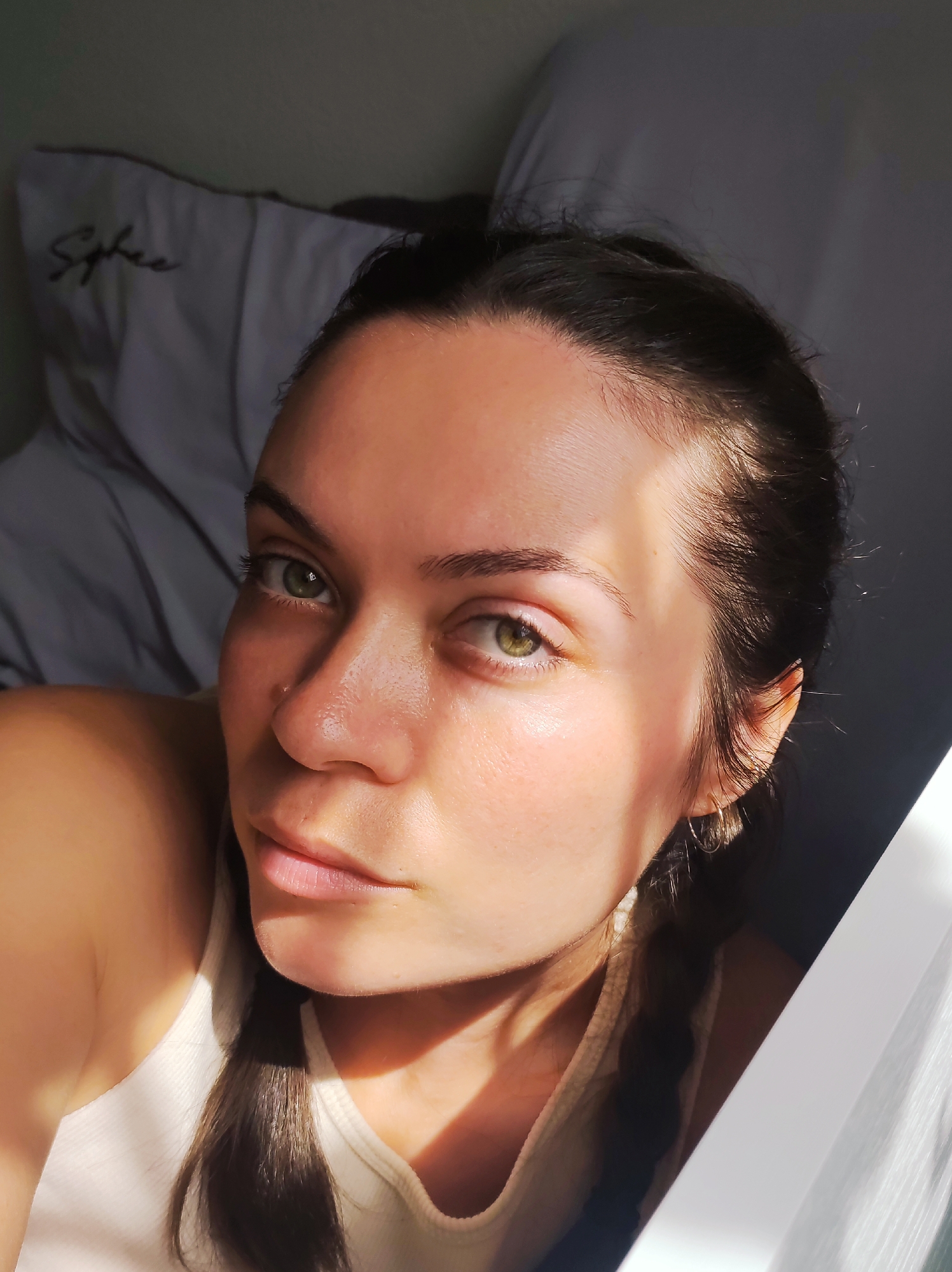 The image is a close-up, high-angle selfie of a young woman with fair skin, green eyes, and dark brown hair. Her hair is styled in a braid that falls over her shoulder. She is wearing a light-colored tank top. The lighting suggests natural sunlight, casting shadows across her face and body. In the background, a pillow with the word "Sphinx" embroidered on it is visible, suggesting the setting is a bedroom. The overall impression is one of a relaxed, intimate moment captured in natural light. The location in the city of Chaikovsky, Russia, is not discernible from the image itself, as there are no visual cues to indicate its geographical context.