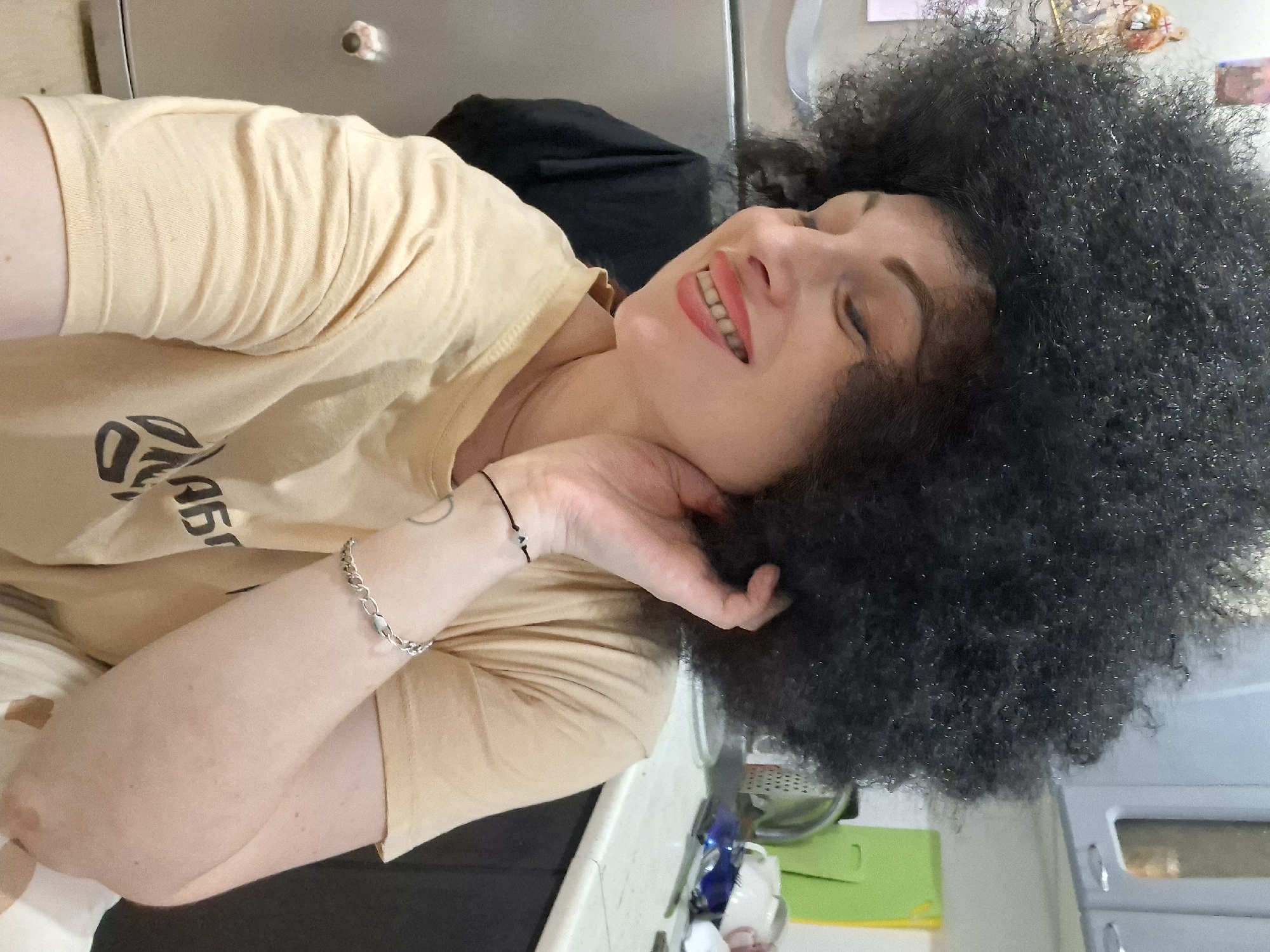 A young woman with voluminous, curly black hair and striking makeup, including bright lipstick and defined eyebrows, is captured in a close-up, self-taken portrait. She is wearing a light tan t-shirt and has a delicate bracelet with silver links and small white beads on her left wrist, as well as a thin black string bracelet with a small bead. She is looking downwards and to her left with a radiant smile, her left hand is touching her neck and hair. The background suggests an indoor setting, possibly a kitchen, with cabinets, a countertop, and appliances visible. The lighting is artificial and casts subtle shadows. No specific location in Myadzyel, Belarus can be determined from the visual information alone.