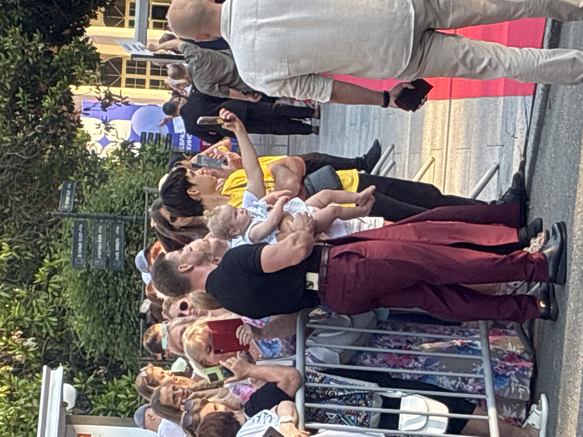 The image shows a crowd of people in what appears to be a public gathering area in Sochi, Russia. In the foreground, a man in a black t-shirt and maroon pants holds a baby. He is surrounded by people, some of whom are taking pictures with their phones. A metal barricade separates the crowd from the street. In the background, there are signs with Russian text, trees, and buildings. The overall scene suggests an event or a celebrity appearance in an outdoor urban setting.