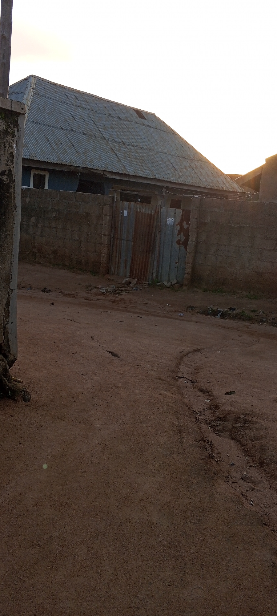 The image depicts an exterior view of a residential building in Jalingo, Nigeria, likely in a semi-urban or rural area. The building features a corrugated metal roof, painted blue on the lower wall sections visible. A concrete block wall encloses the property, with a corrugated metal gate that appears weathered and rusty. The ground in front of the property is unpaved and appears to be a dirt road or path. The sky is visible in the upper portion of the image and appears to be overcast with the setting sun creating a slight glow. There are no people or animals clearly visible in the image. The overall scene suggests a simple, functional dwelling.