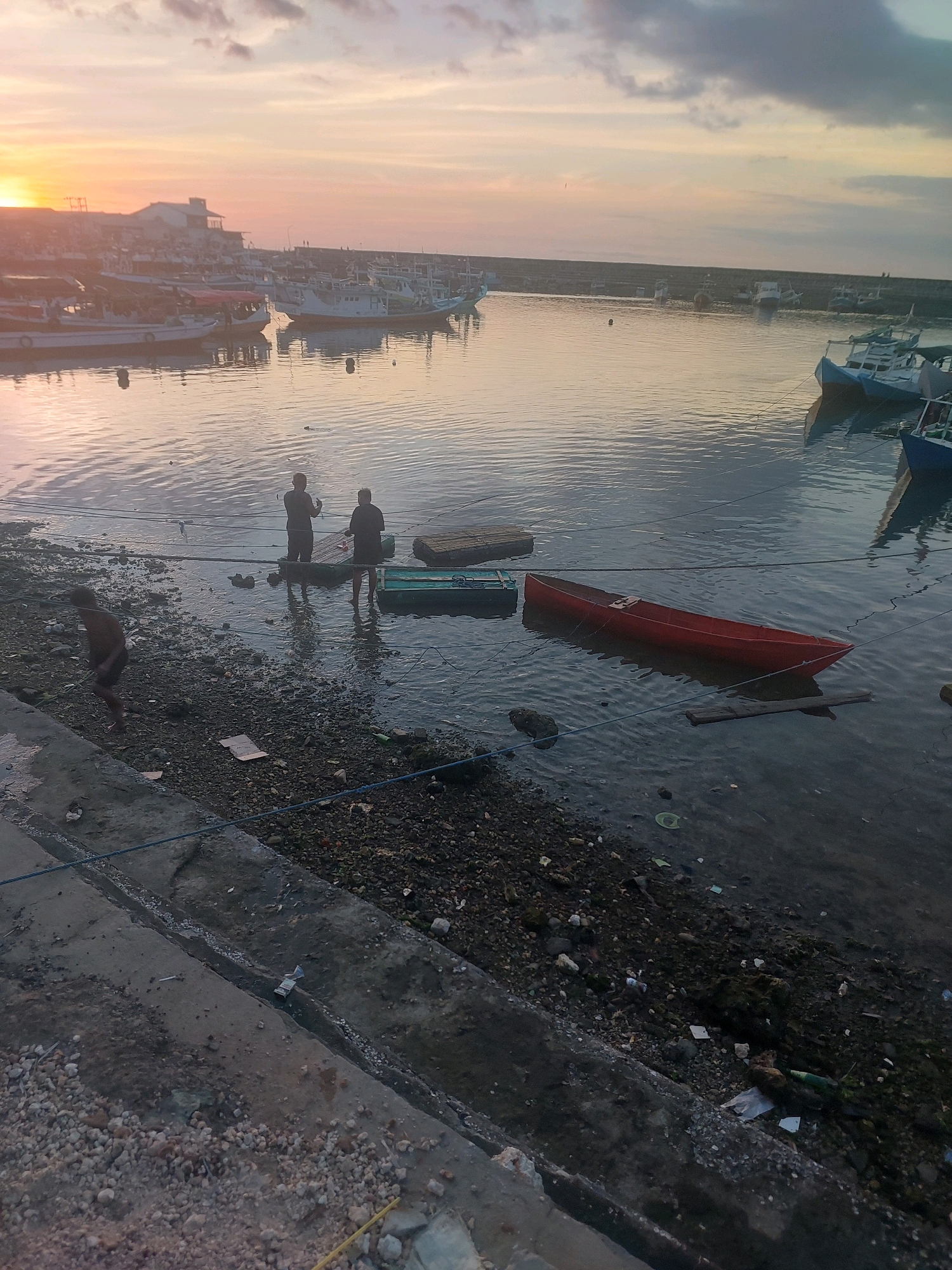 This is a photograph taken at dusk, likely in the city of Kupang, Indonesia. The scene depicts a harbor or bay area with several boats anchored in the water. The sky is a mix of soft pinks, oranges, and grays, indicating the setting sun.
In the foreground, a person, appearing to be a young boy, is walking along a concrete embankment which is littered with rocks and debris. He is facing away from the camera and moving towards the left of the frame.
Further into the water, closer to the shore, three figures are visible standing in the shallow water. Two of them are adult males, one facing the other, and the third figure is partially obscured. They appear to be engaged in an activity, possibly fishing or attending to the boats. There are two small boats near them, one green and one red, the red one being a prominent subject in the middle ground. The red boat is empty and appears to be a simple, wooden vessel.
In the background, numerous fishing boats are moored in rows, suggesting a working harbor. Buildings, possibly part of a port or town, are visible along the distant shoreline. The water is relatively calm, reflecting the colors of the sky. The overall atmosphere is one of a typical evening in a coastal Indonesian community. The presence of scattered litter along the shoreline suggests an environment that is not meticulously maintained.