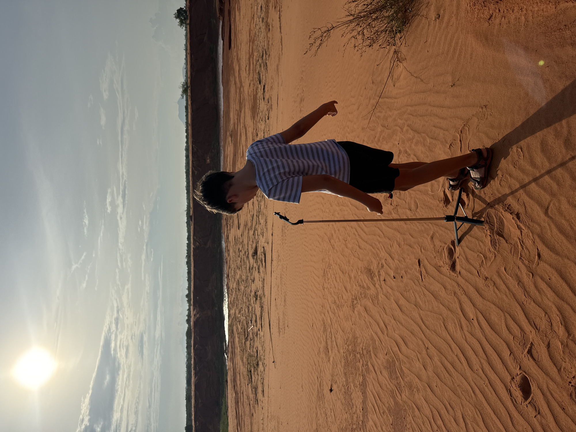 In Mui Ne, Vietnam, a young boy stands on a sandy terrain with rippled sand dunes. He is wearing a striped t-shirt and black shorts, with sandals on his feet. The boy is looking down at a small tripod with a phone attached to it. In the background, there is a body of water, likely a river or lake, with vegetation on the opposite bank. Further in the distance, a mountain range can be seen. The sky is partly cloudy with the sun shining brightly, casting a shadow of the boy and the tripod on the sand. The overall scene depicts a boy engaging with technology in a natural, outdoor setting.