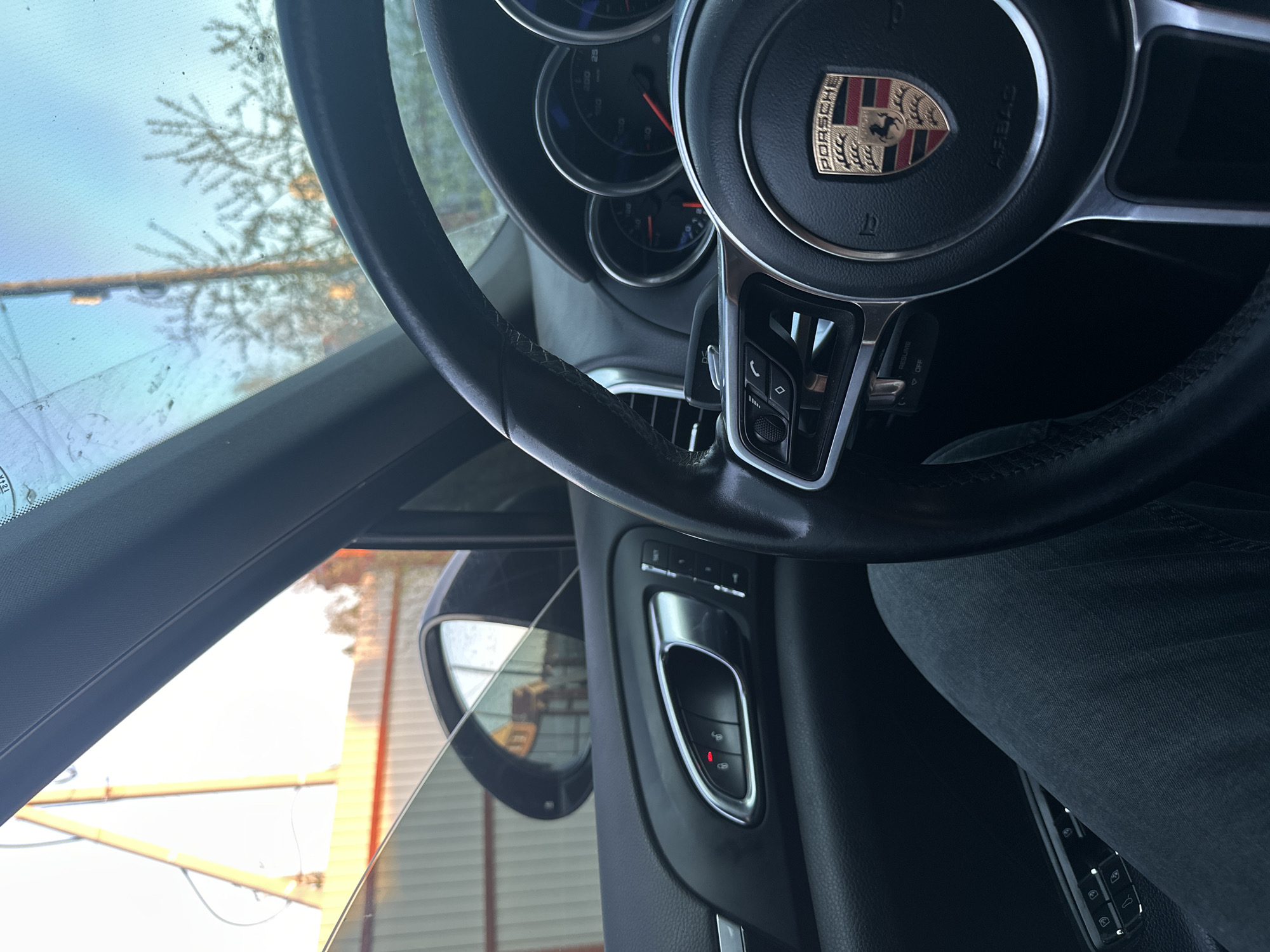 The image shows the interior of a Porsche car, focusing on the steering wheel and the driver's side door panel. The Porsche logo is prominent in the center of the steering wheel. The door panel features the window and mirror controls. Outside the window, a blurred view of a building under construction is visible. The person is wearing dark jeans. The image does not provide information about the location in the city of Unknown, Unknown.