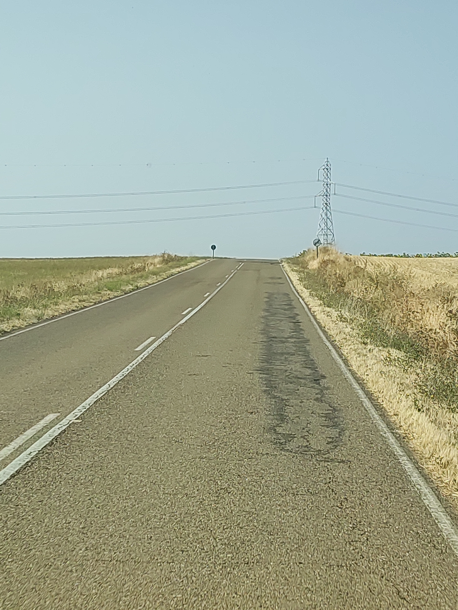 The image depicts a straight, asphalt road leading towards a slight incline under a clear, pale blue sky. The road is marked with white lines, with a dashed line separating lanes and solid white lines along the edges. To the left of the road, a dry, grassy field extends to the horizon. To the right, a harvested field, likely of wheat or barley, is visible, with dry stalks and some green vegetation. In the distance, a tall, lattice-style electricity pylon stands against the sky, with power lines extending horizontally from it. A circular blue traffic sign is positioned on the right side of the road, atop a short post, near the crest of the incline. The scene appears to be a rural landscape in Baquerín de Campos, Spain, suggesting a remote, open area. There are no people or vehicles visible in the image. The overall impression is one of a quiet, sun-drenched day in the Spanish countryside.