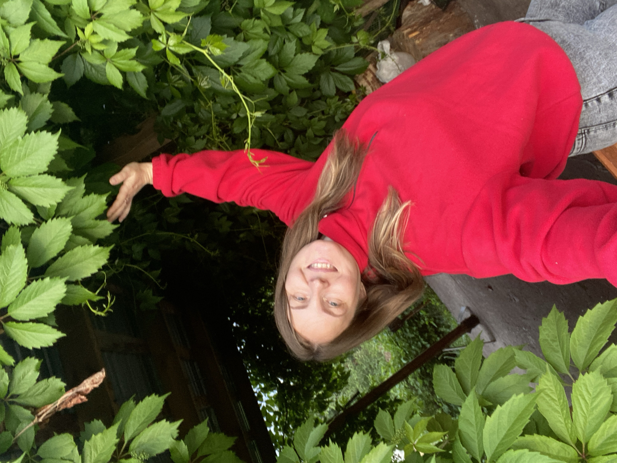 A young woman with long, light brown hair and blue eyes smiles directly at the camera in a tilted outdoor selfie. She is wearing a vibrant red long-sleeved high-neck top or sweater and light grey jeans, visible at the bottom right. Her left arm is raised and extended upwards, with her open hand reaching towards a dense canopy of bright green, multi-lobed leaves, likely from a climbing vine. A small ring is visible on the fourth finger of her left hand. The background is lush with similar green foliage. Through the leaves in the upper left, dark wooden beams or structural elements of what appears to be a pergola or arbor are visible. A small, dry brown leaf or twig hangs from the foliage near the wooden structure in the upper left. The lower right portion of the image reveals a dark ground surface, possibly concrete, with some lighter-colored debris. The scene is illuminated by natural daylight.