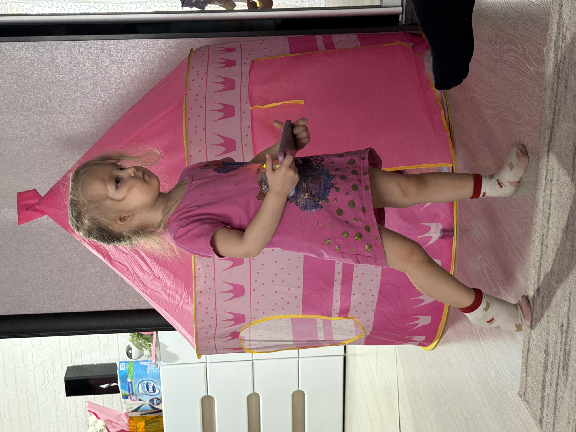 A young child, likely a girl with blonde hair, stands upright in an indoor setting. She is wearing a short-sleeved pink dress adorned with gold polka dots and a sequined design on the front. Her white socks, featuring red cuffs and small patterns (possibly bunnies), are visible on her feet. The child holds a dark-colored smartphone in both hands, gazing intently towards the left of the frame.
To her right is a vibrant pink play tent, styled like a castle or teepee, with patterns of white polka dots and yellow crowns along its base and top. Part of an adult's leg, clad in dark pants, is visible in the upper right background, next to a light-colored rug or carpet.
The background reveals a domestic environment with a light-patterned wall or wallpaper and white furniture or shelving. Household items are discernible on a shelf behind the tent, including packages of toilet paper (one clearly showing the brand "Clean" and "Light") and a dark box. The lighting suggests daytime indoors.