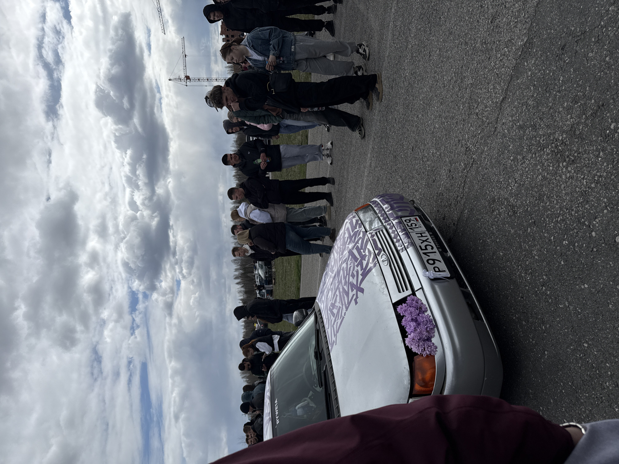 A line of people stand along the edge of an asphalt surface, with a light-colored car decorated with purple flowers parked nearby. The license plate on the car reads "P915XH 159 RUS," indicating registration in Russia. In the background, construction cranes are visible against a cloudy sky. The location is likely Perm, Russia, based on the license plate.