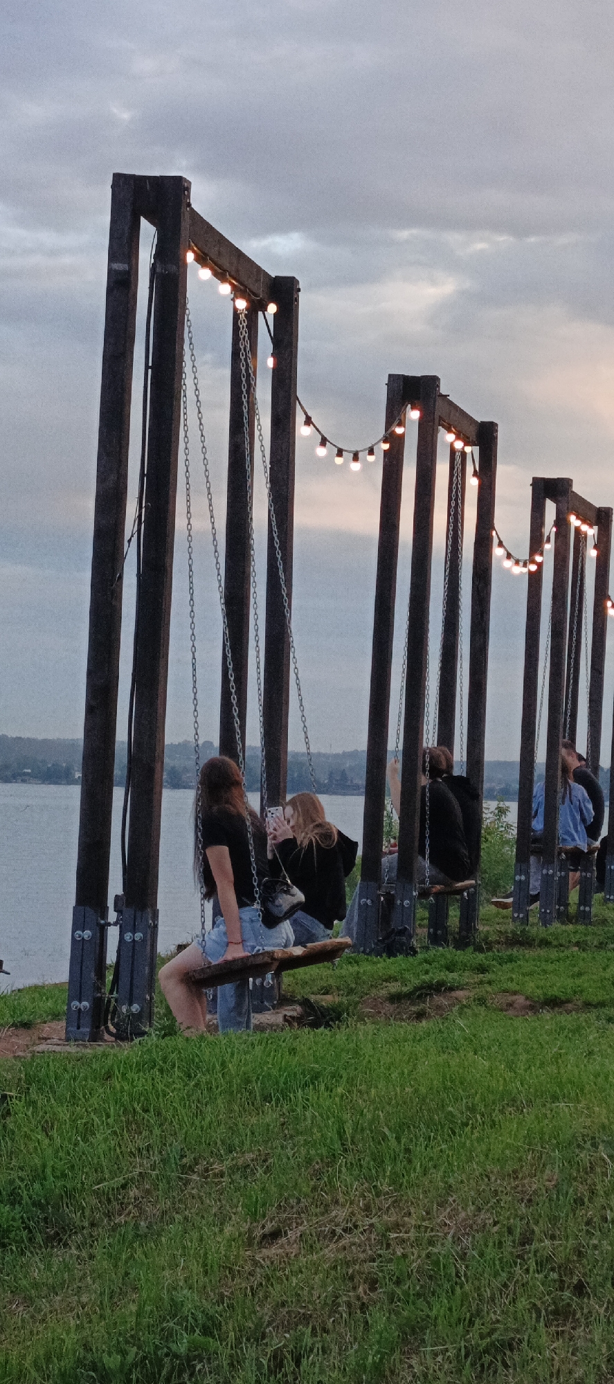 The image depicts a scene at a recreational area along the shore of Lake Votkinsk in Votkinsk, Russia. Several wooden swing structures, each with chains and a swing seat, are lined up. String lights are illuminated above the swings, casting a warm glow against the cloudy twilight sky. In the foreground, lush green grass covers the slope leading down to the lake.
On one of the swings, two young women are seated. The woman on the left, with long dark hair, is wearing a black t-shirt and denim shorts. She is looking towards the right, and her body is positioned to face the swing. The woman on the right, with lighter hair, is wearing a black hoodie and is holding a smartphone, appearing to take a picture or video. Her gaze is directed at the phone.
Further down the line of swings, other individuals are visible, though less clearly. One person is sitting on a swing, and another person in the distance is partially obscured. The calm water of Lake Votkinsk stretches out behind the swing structures, with a distant shoreline visible. The overall atmosphere is serene and relaxed, suggesting an evening outing.